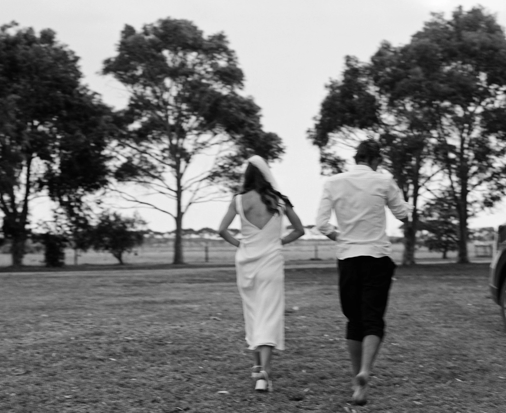 Black and white photo of a woman in a white dress and a man in dark shorts walking away through a park with trees in the background.