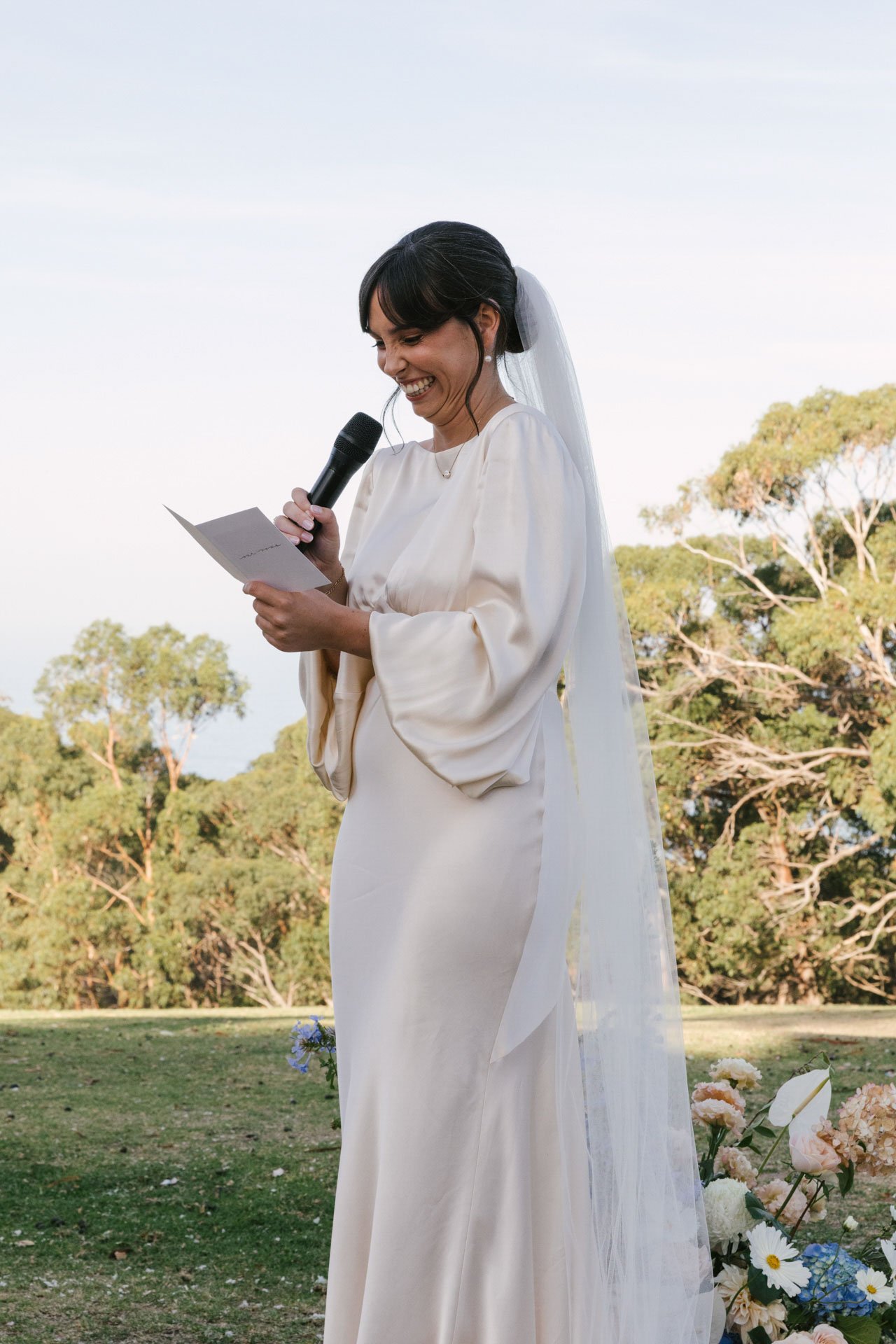 A woman in a white wedding dress and veil holding a microphone and reading from a piece of paper, standing outdoors with trees and flowers in the background.