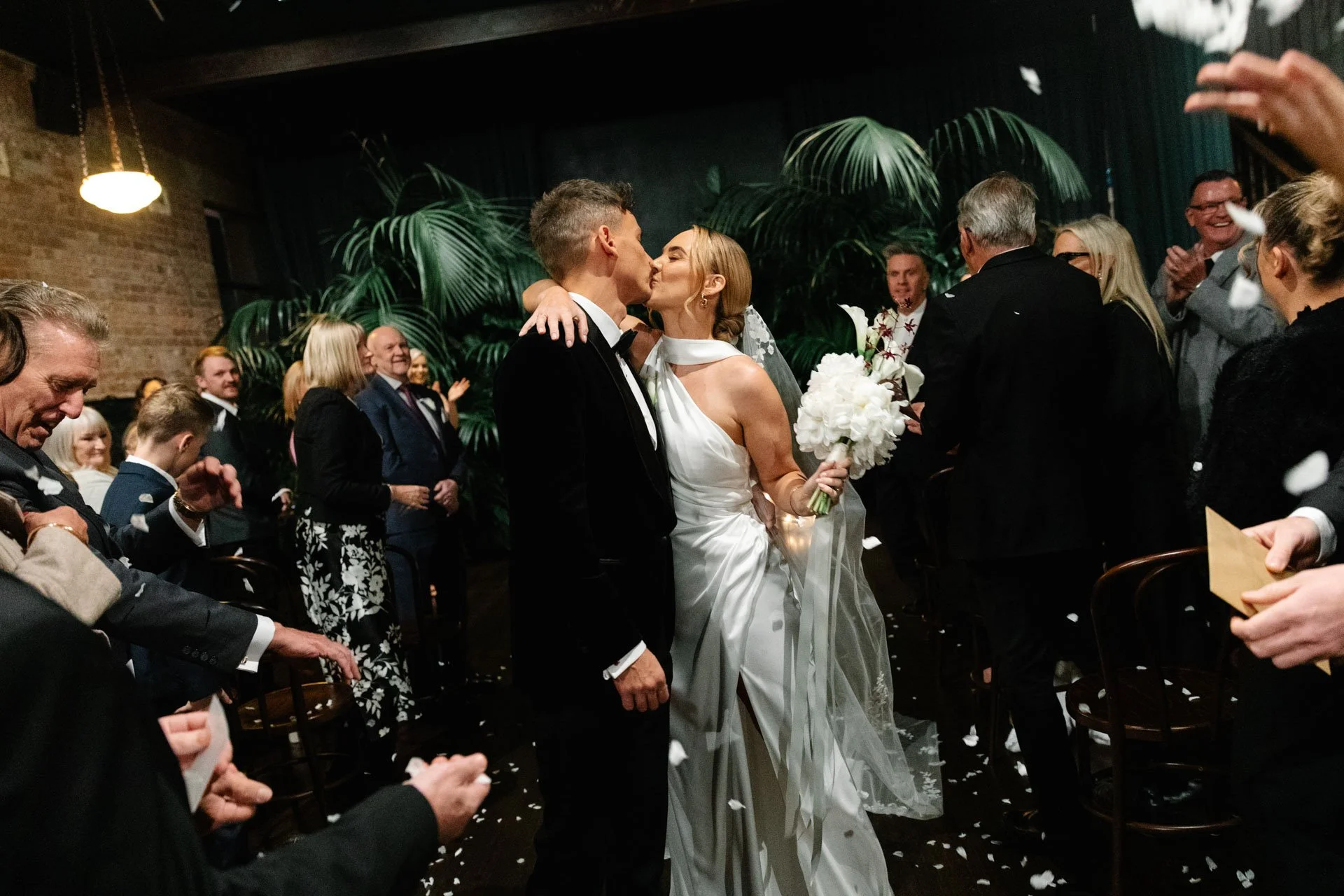 A bride and groom kiss during their wedding ceremony, surrounded by guests in an indoor venue decorated with large green plants and dark curtains.