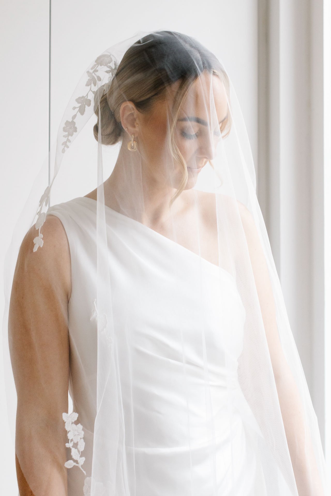 Bride with blonde hair wearing a white sleeveless dress and veil, standing indoors near a window with light-colored curtains.