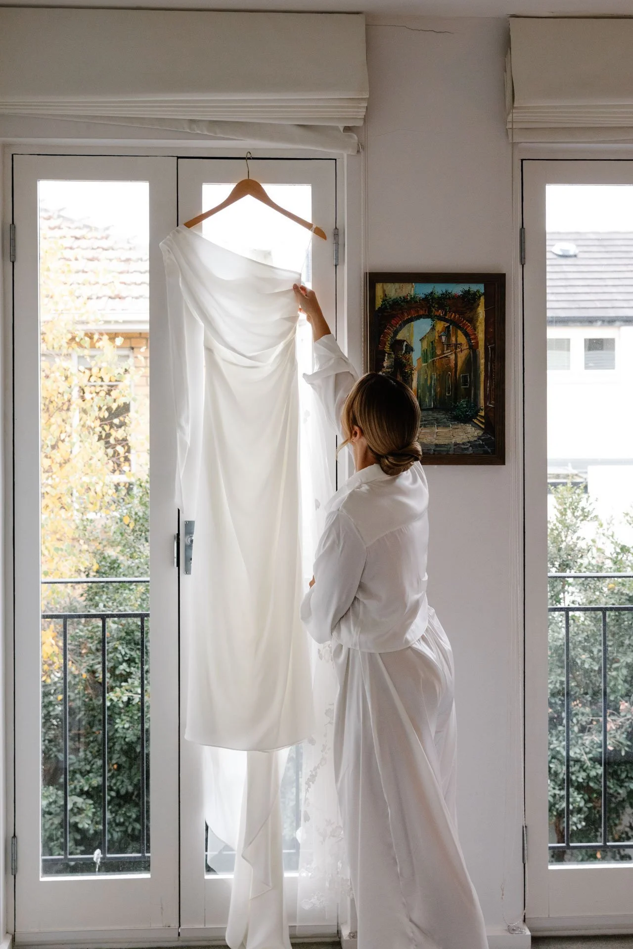 A woman in white pajamas holding a white dress on a hanger in front of glass doors in a bright room.