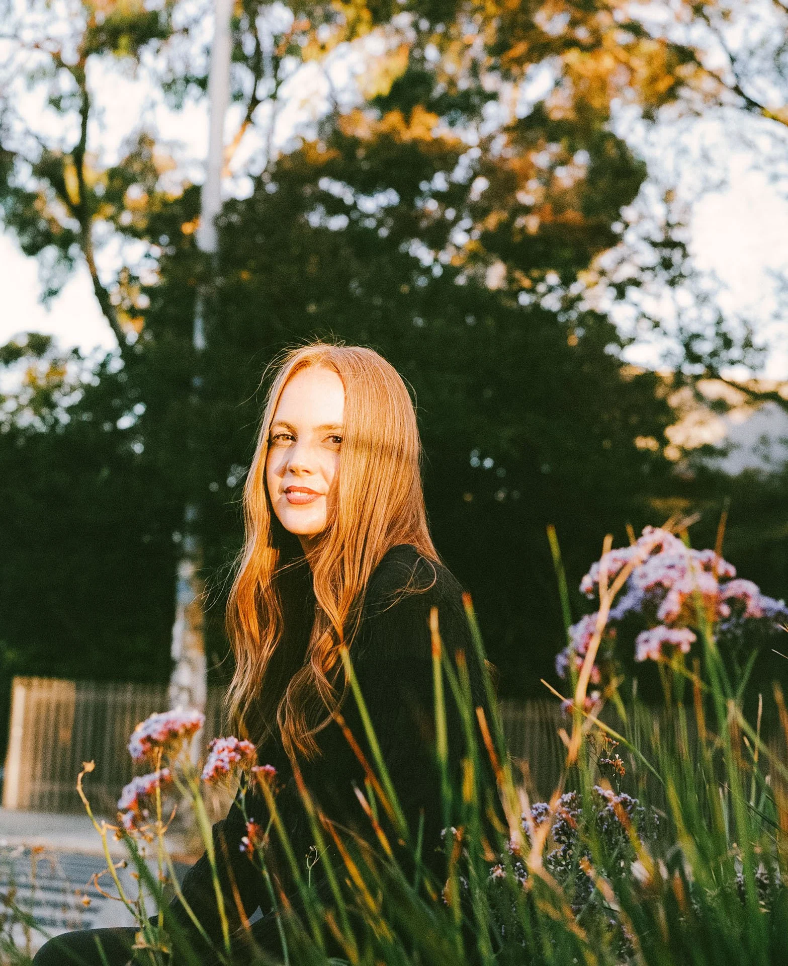 A young woman with long red hair smiling outdoors in front of trees and flowers, captured during golden hour