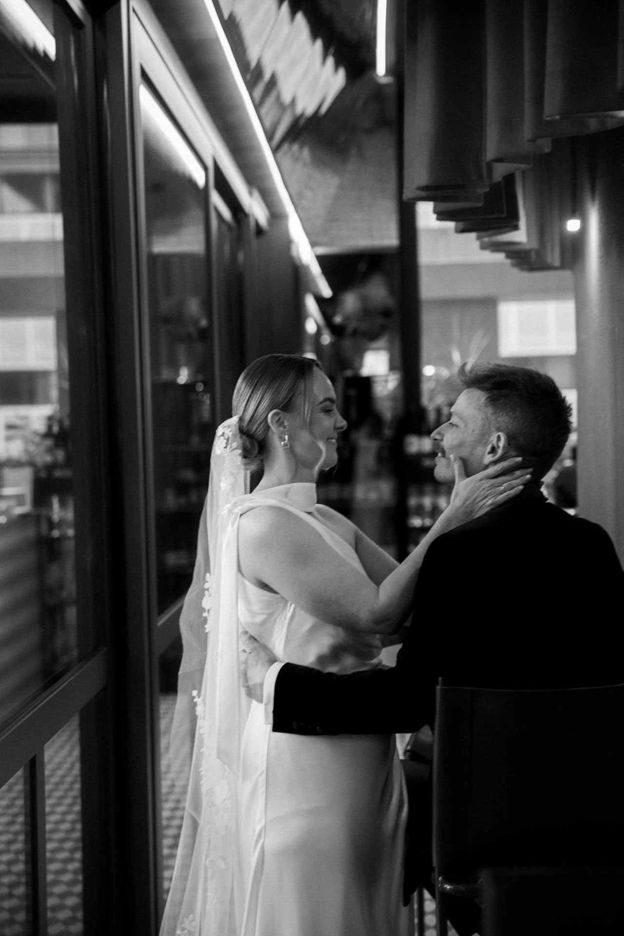 A bride and groom share an intimate moment, facing each other closely, with the bride smiling and the groom holding her face, inside a venue with large glass windows.