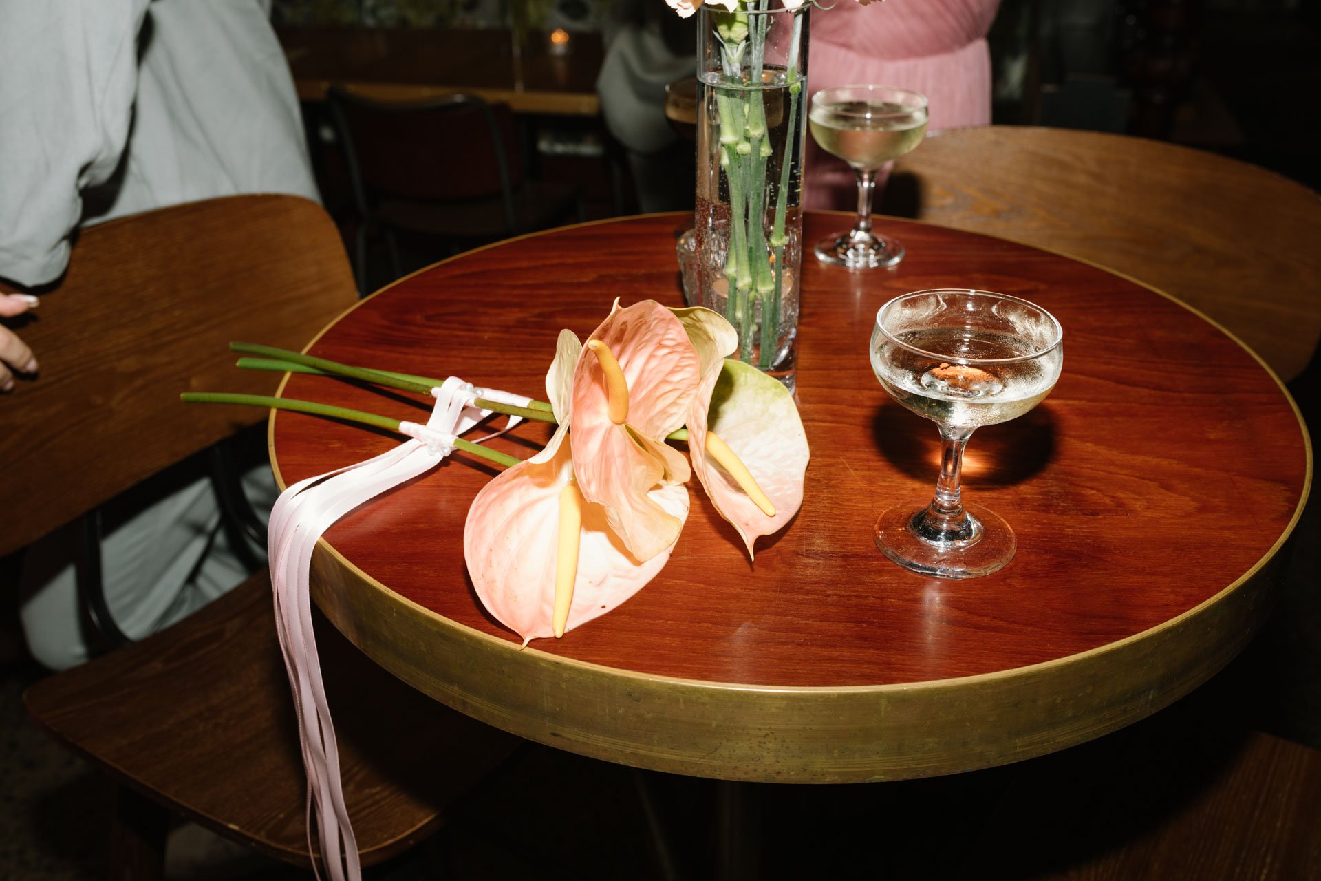 A round wooden table decorated with a pink anthurium flower, a tall glass vase with green flower stems, two coupe glasses with white wine, and other floral arrangements in the background.