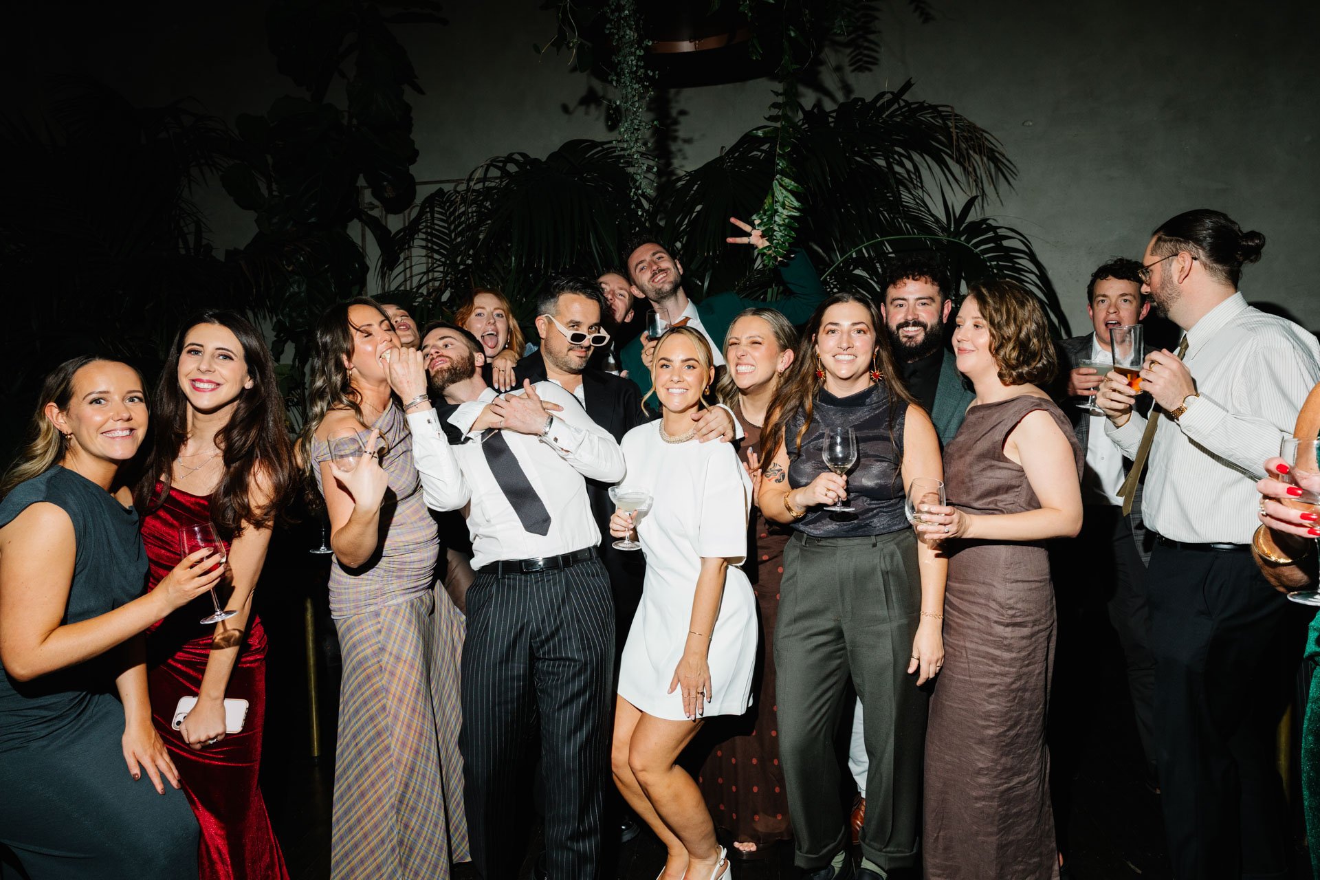 A group of people dressed in semi-formal attire at a social gathering, holding drinks and smiling together.