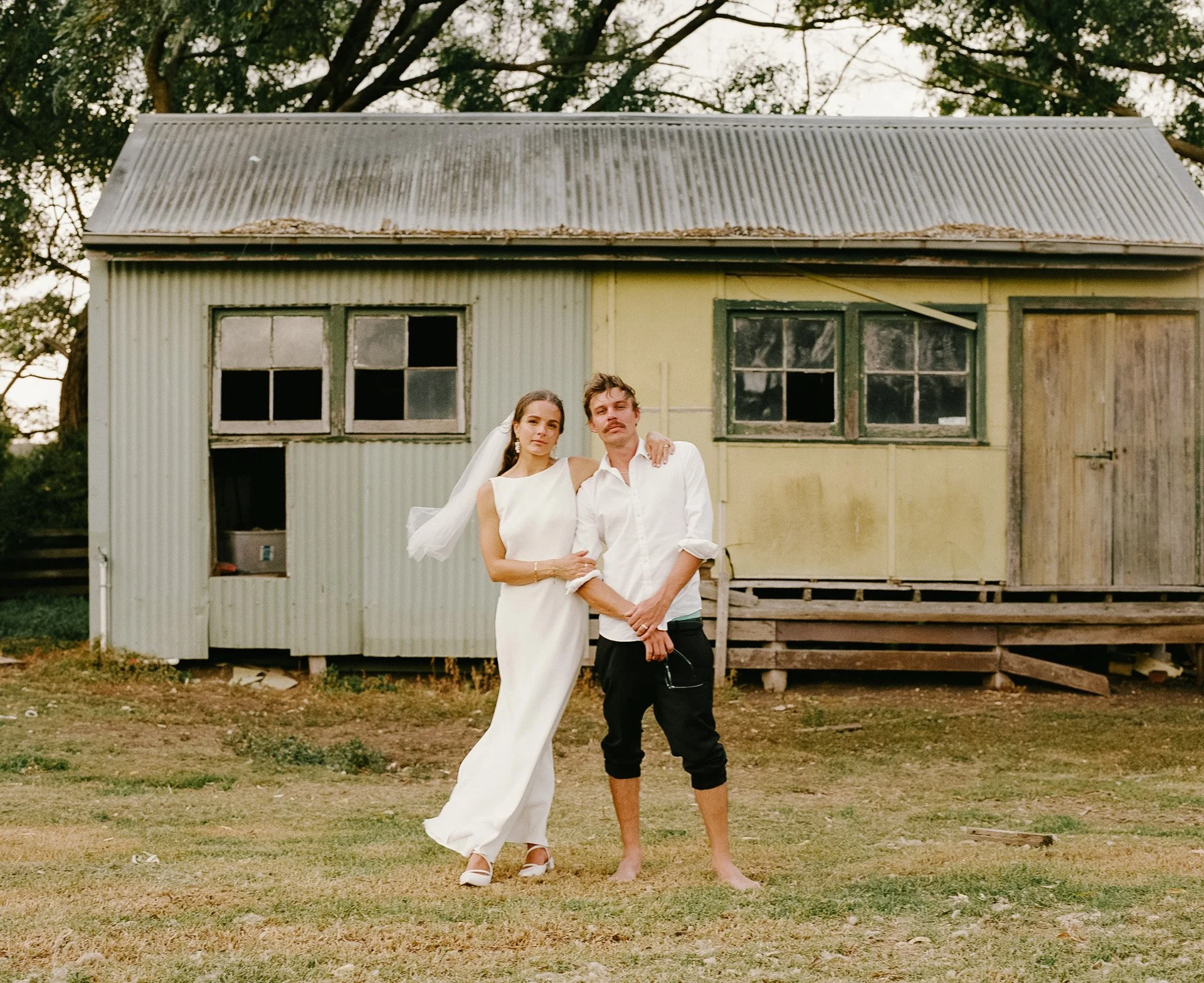 A couple, a woman in a wedding dress and a man in a white shirt, standing outdoors in front of a weathered, old house with peeling paint and broken windows. The woman has her arm around the man's shoulder, and they are posing for a photo.