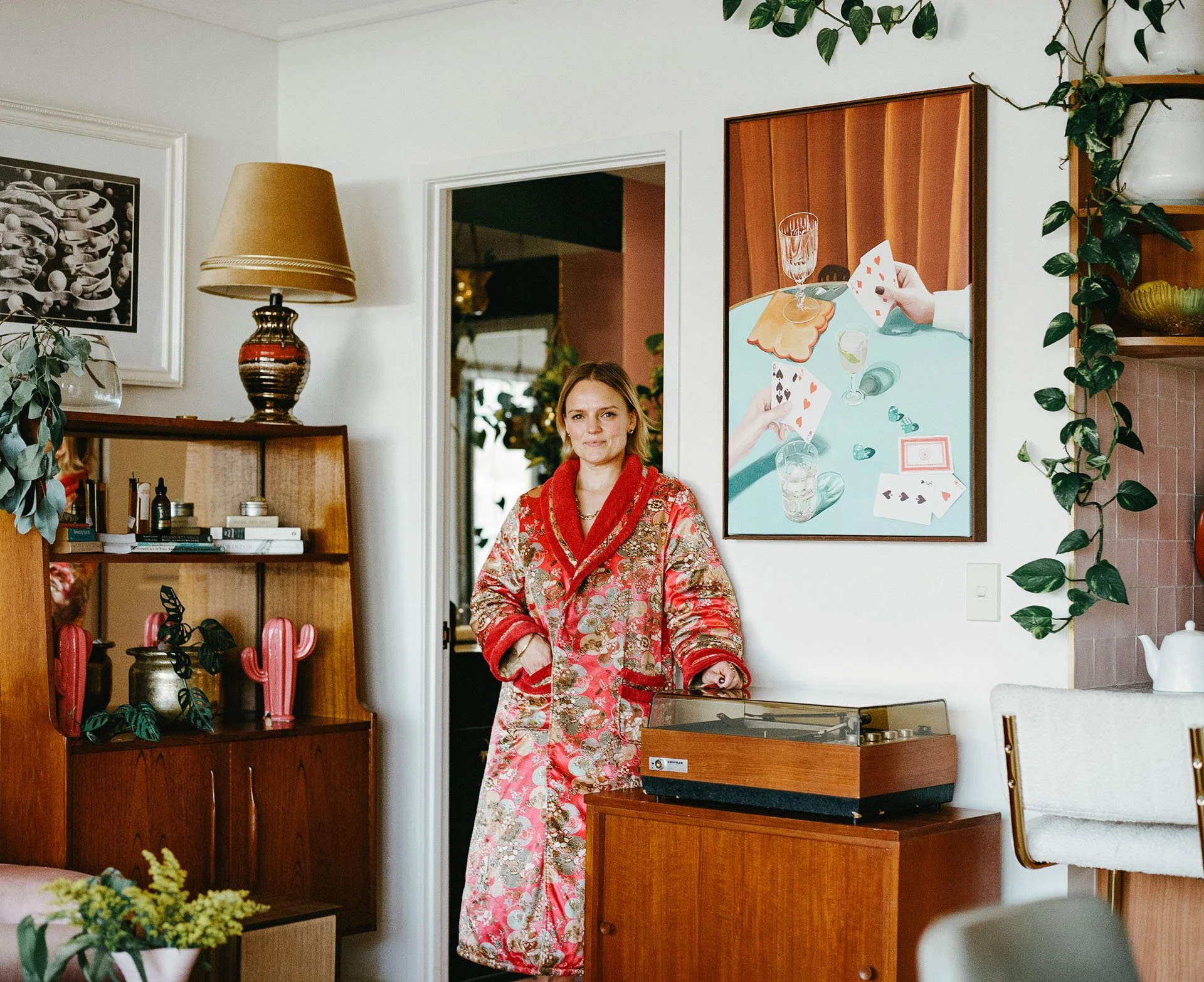 A woman in a red, floral patterned robe standing in a living room, with a vintage record player on a wooden cabinet nearby, and various houseplants and artwork decorating the space.
