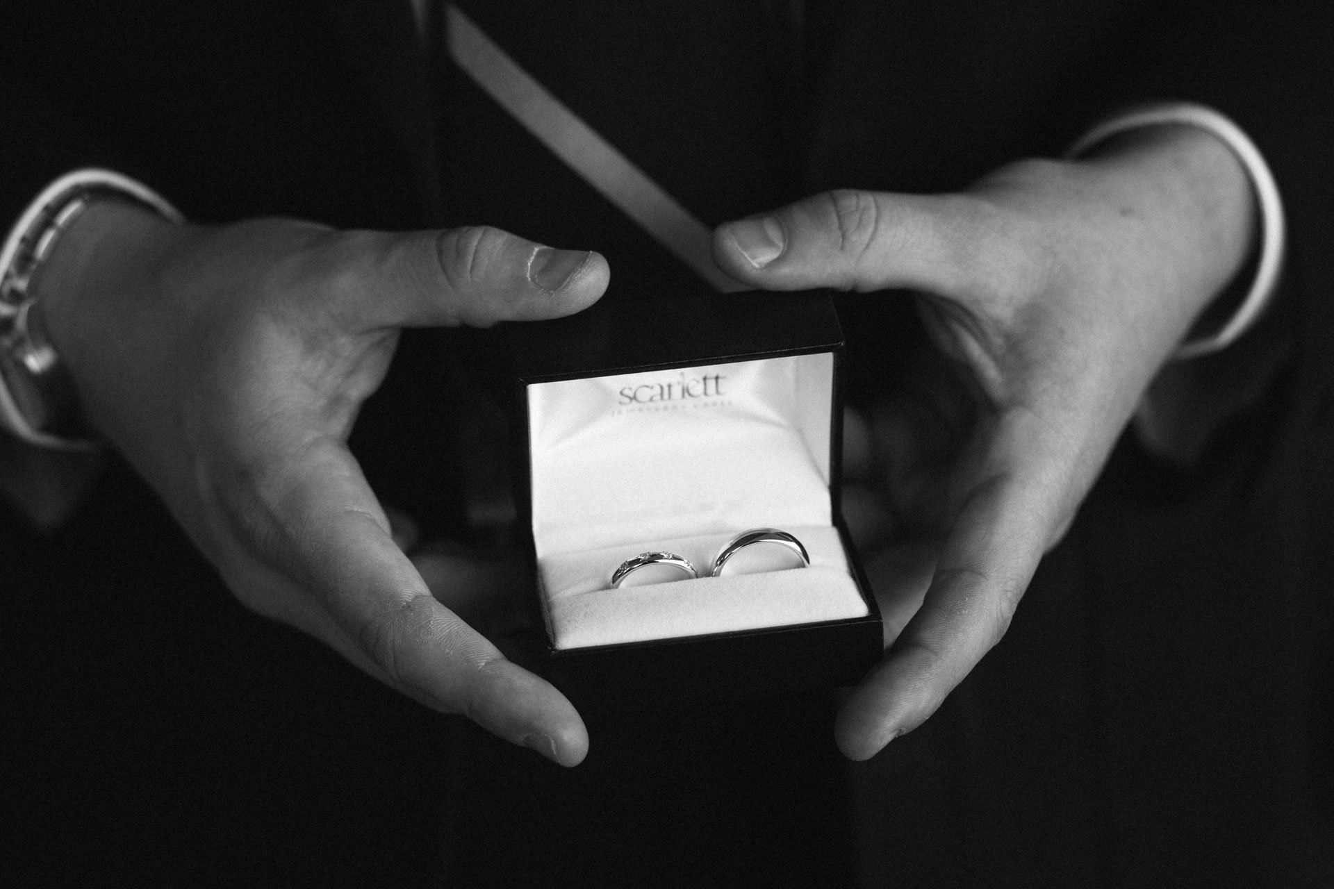 A person holding a jewelry box with two wedding rings inside, on a dark surface.