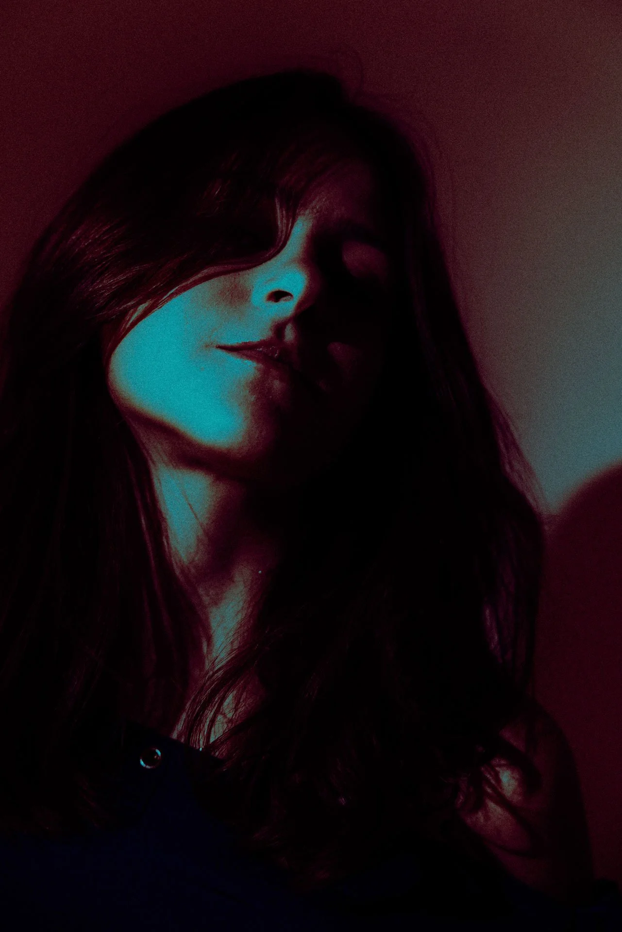 A woman with long dark hair and a slight smile, partially obscured by shadow and colorful lighting.