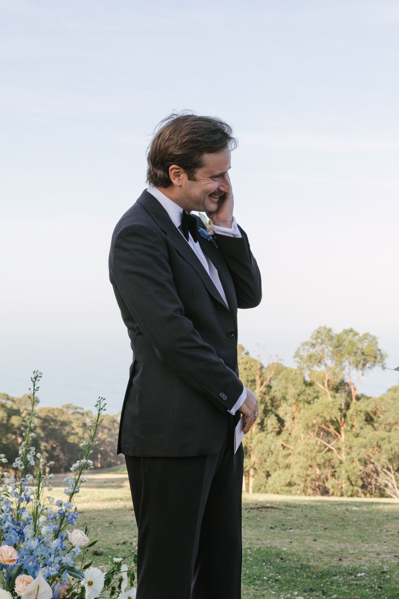 A man in a tuxedo smiling and holding his hand, standing outdoors near a floral arrangement, with trees and a clear sky in the background.