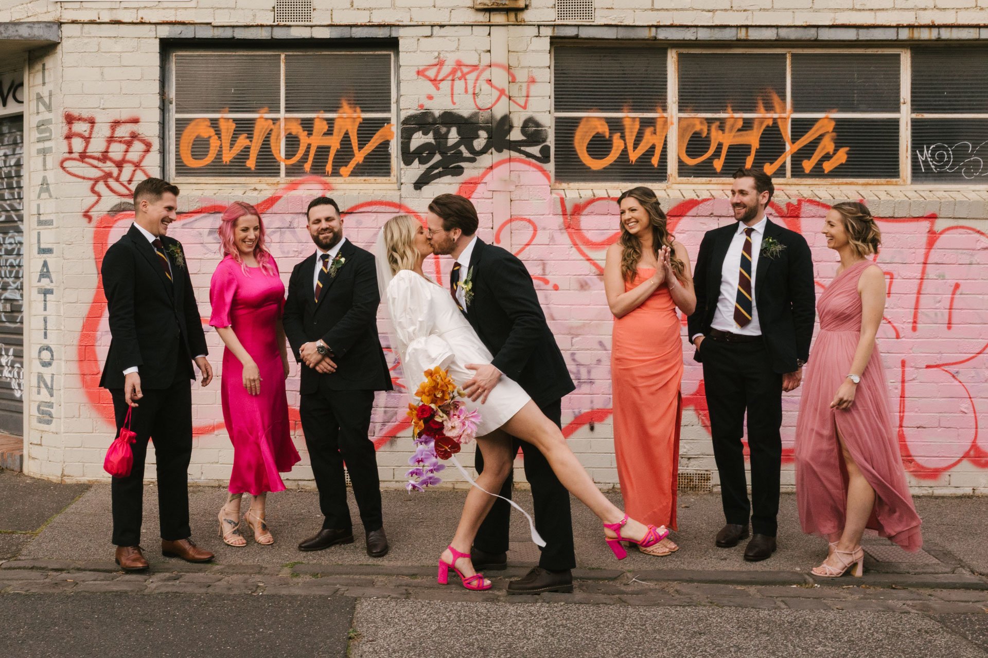 A group of seven people, dressed formally with women in colorful dresses and men in suits, standing in front of a graffiti-covered wall. Two of the women and one of the men are kissing, while the others are smiling and looking at each other.