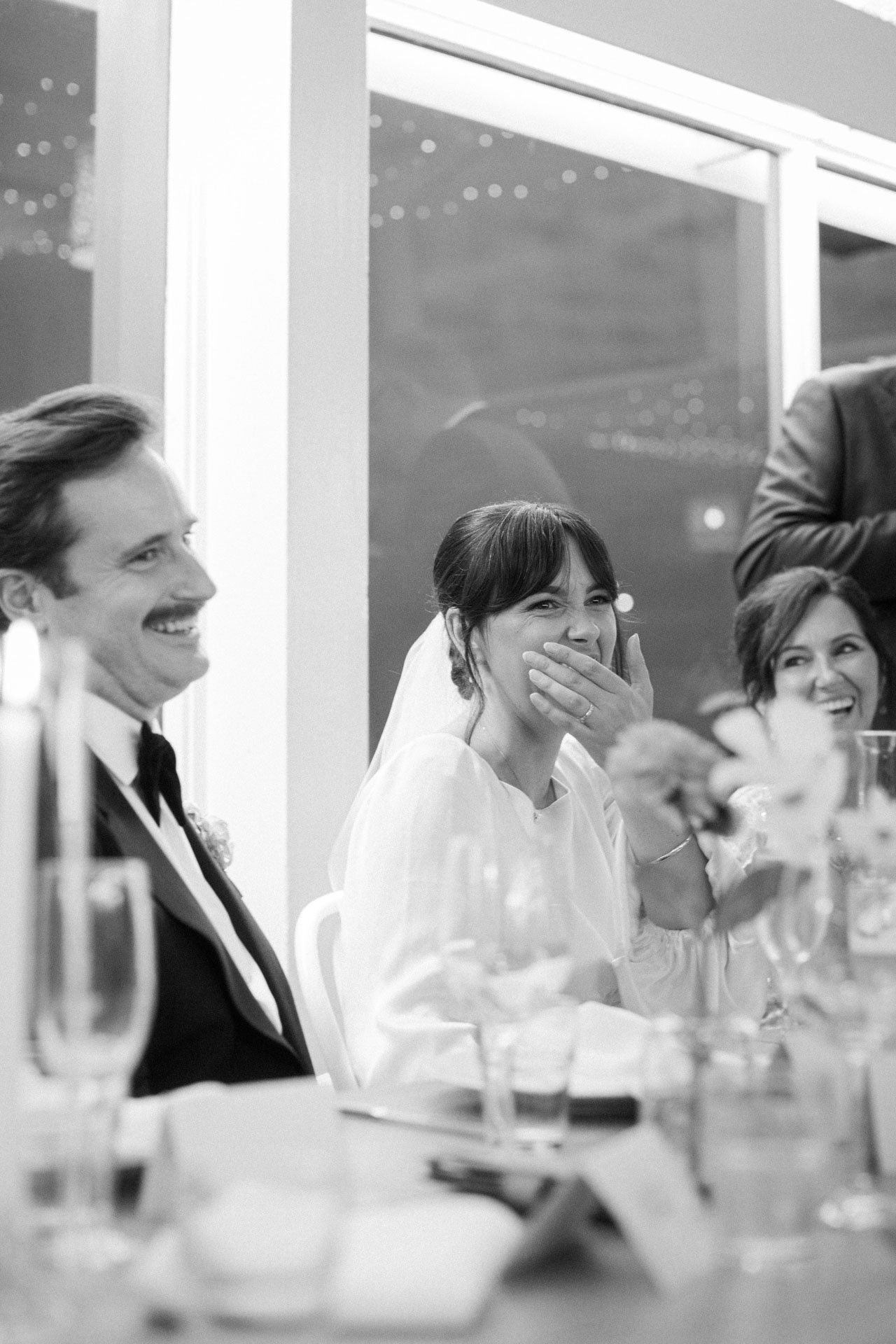 Black and white photo of people at a formal event, sitting at a dinner table with glasses and plates. A woman with dark hair and a white outfit is laughing and covering her mouth. A woman with light hair and a dark suit is smiling on the left, and an