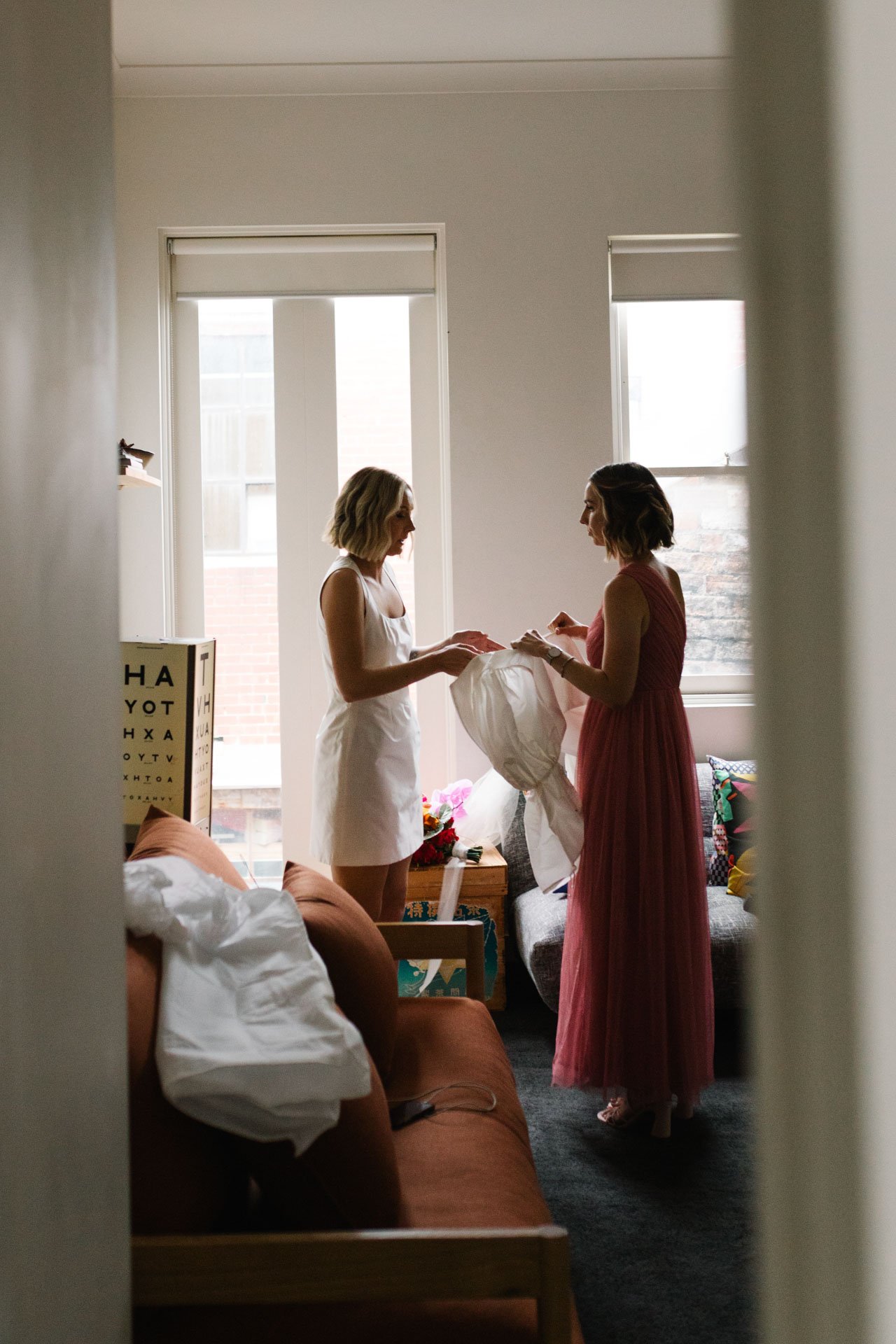 Two women with shoulder-length hair, one in a white dress and the other in a pink dress, are standing and talking inside a brightly lit room with large windows. One woman is holding a white fabric or dress. The room is furnished with a sofa, pillows,