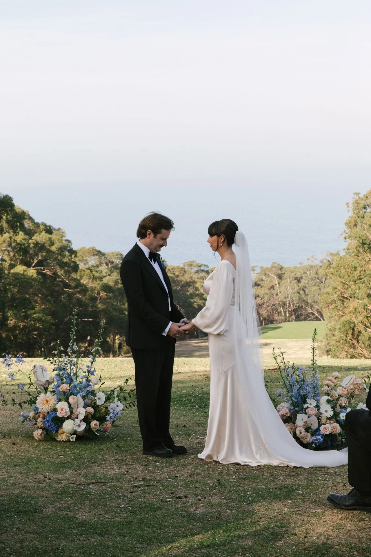 A bride and groom holding hands during their outdoor wedding ceremony, with floral arrangements and trees in the background.