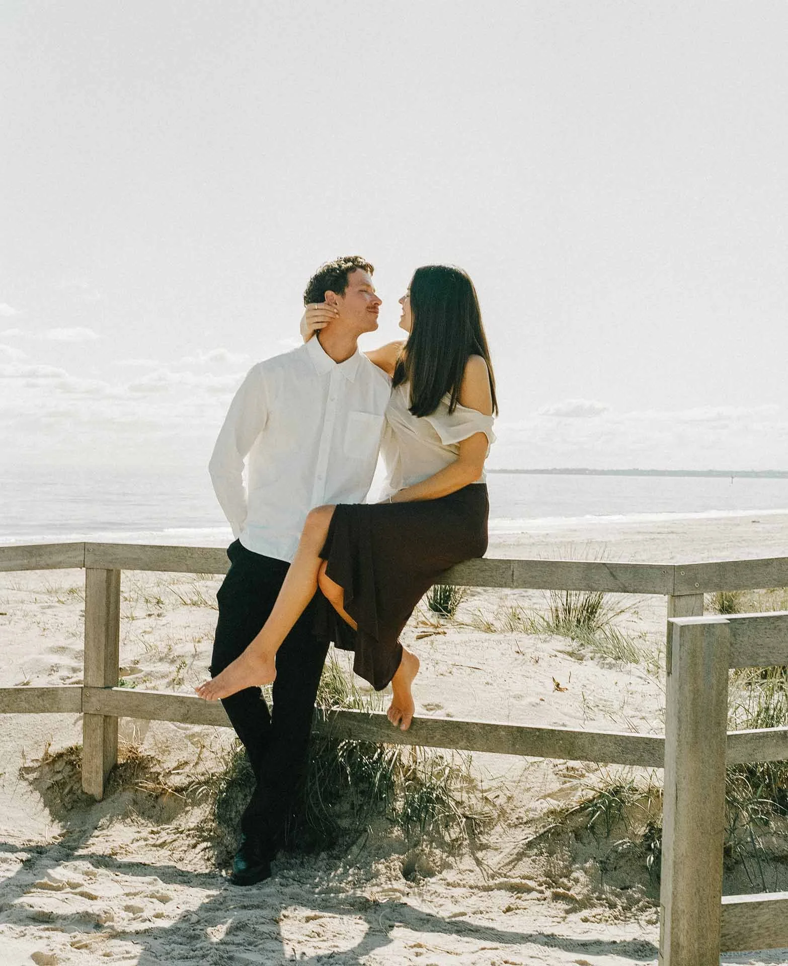 A man and woman at the beach, the man in a white shirt and the woman in a black skirt and white top, standing behind a sand fence with the ocean in the background.