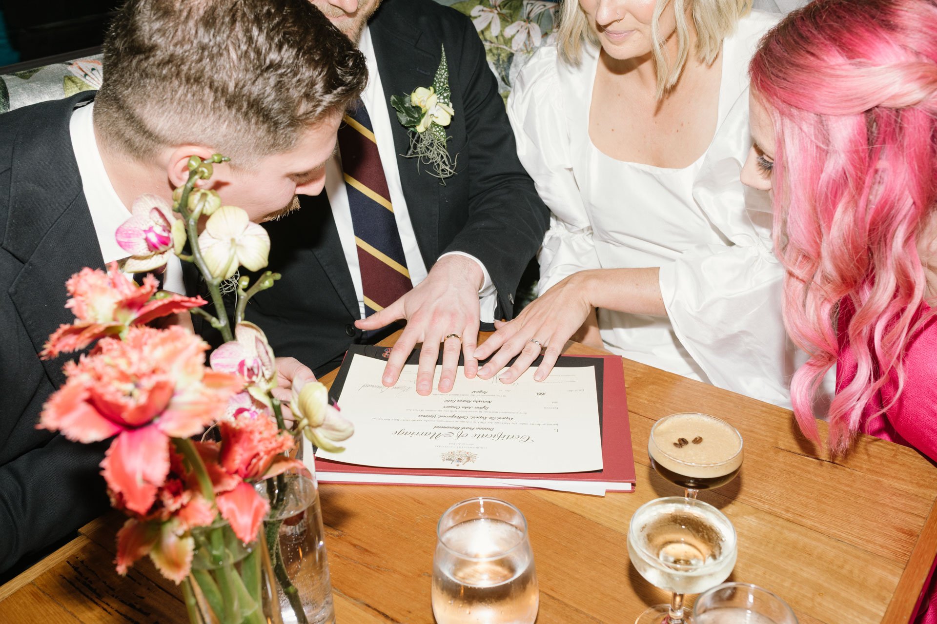 Four people, dressed in formal attire, gather around a table during a wedding ceremony, with a flower arrangement and glasses of champagne visible.