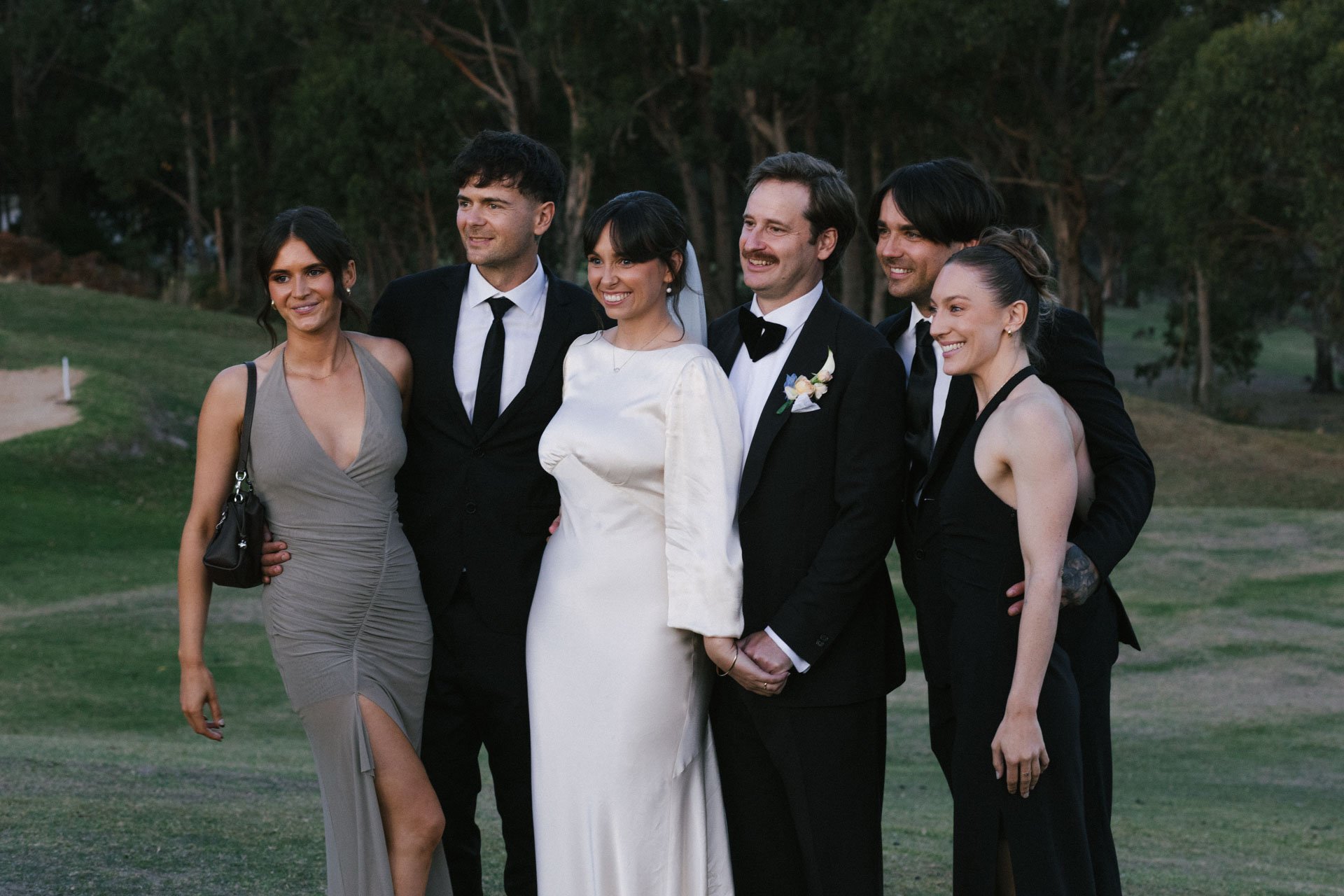 Group of six people at a wedding or formal event outdoors, dressed in elegant attire, smiling and posing together with a wooded background.