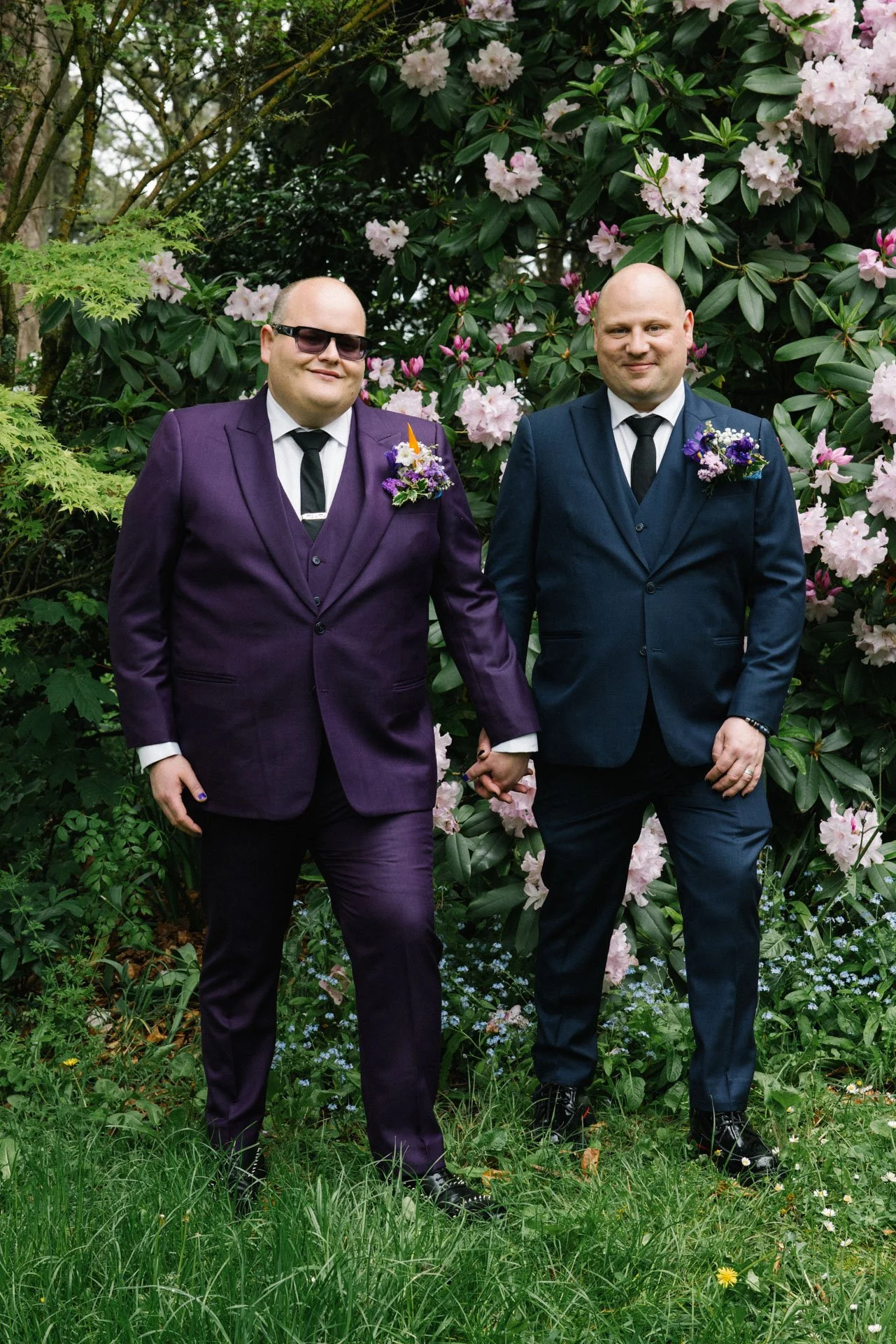Two men dressed in suits, holding hands, standing in a garden with large blooming pink flowers and greenery.
