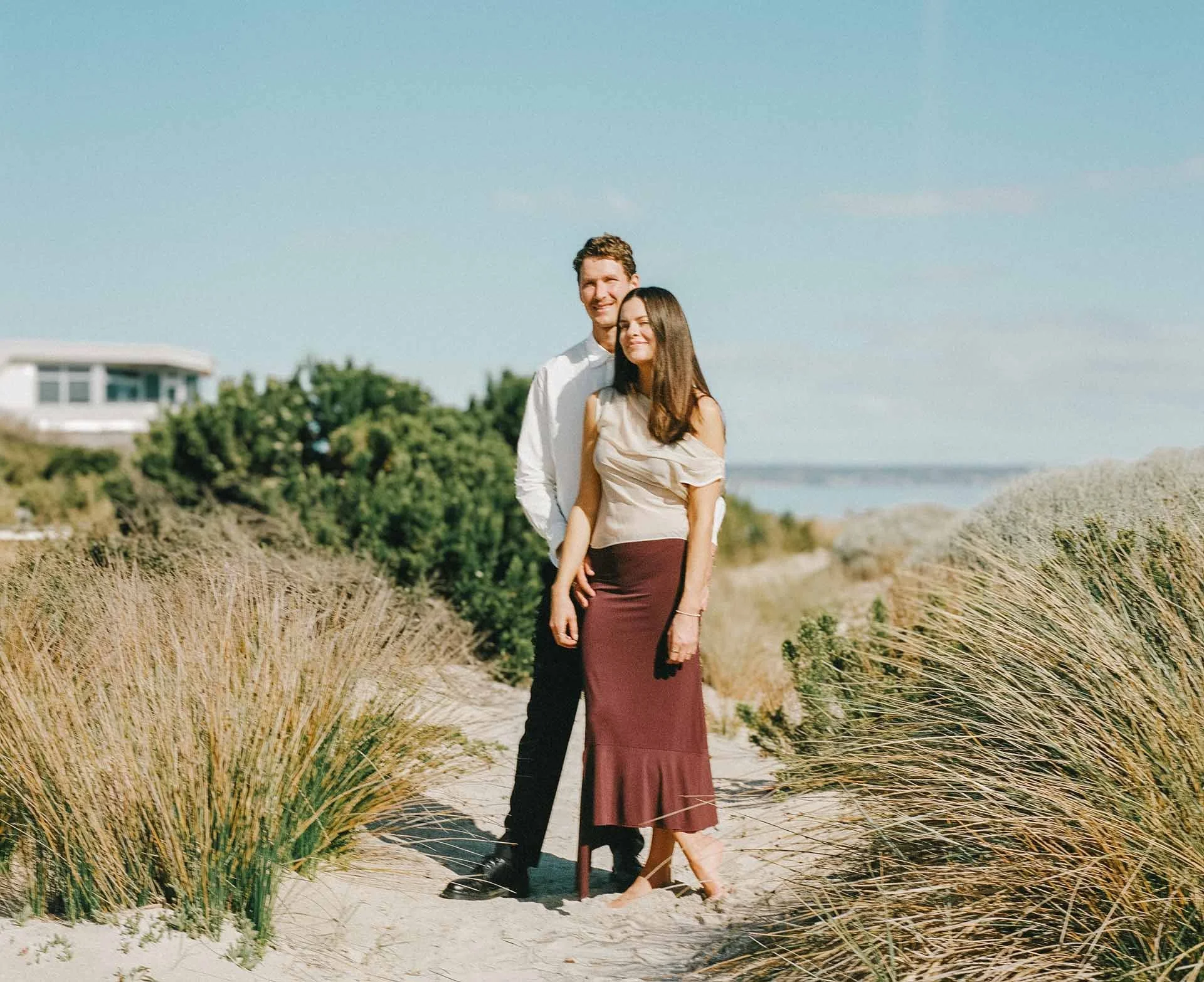 A man and woman standing on a sandy trail surrounded by tall grasses, with a beach and house visible in the background under a clear blue sky.