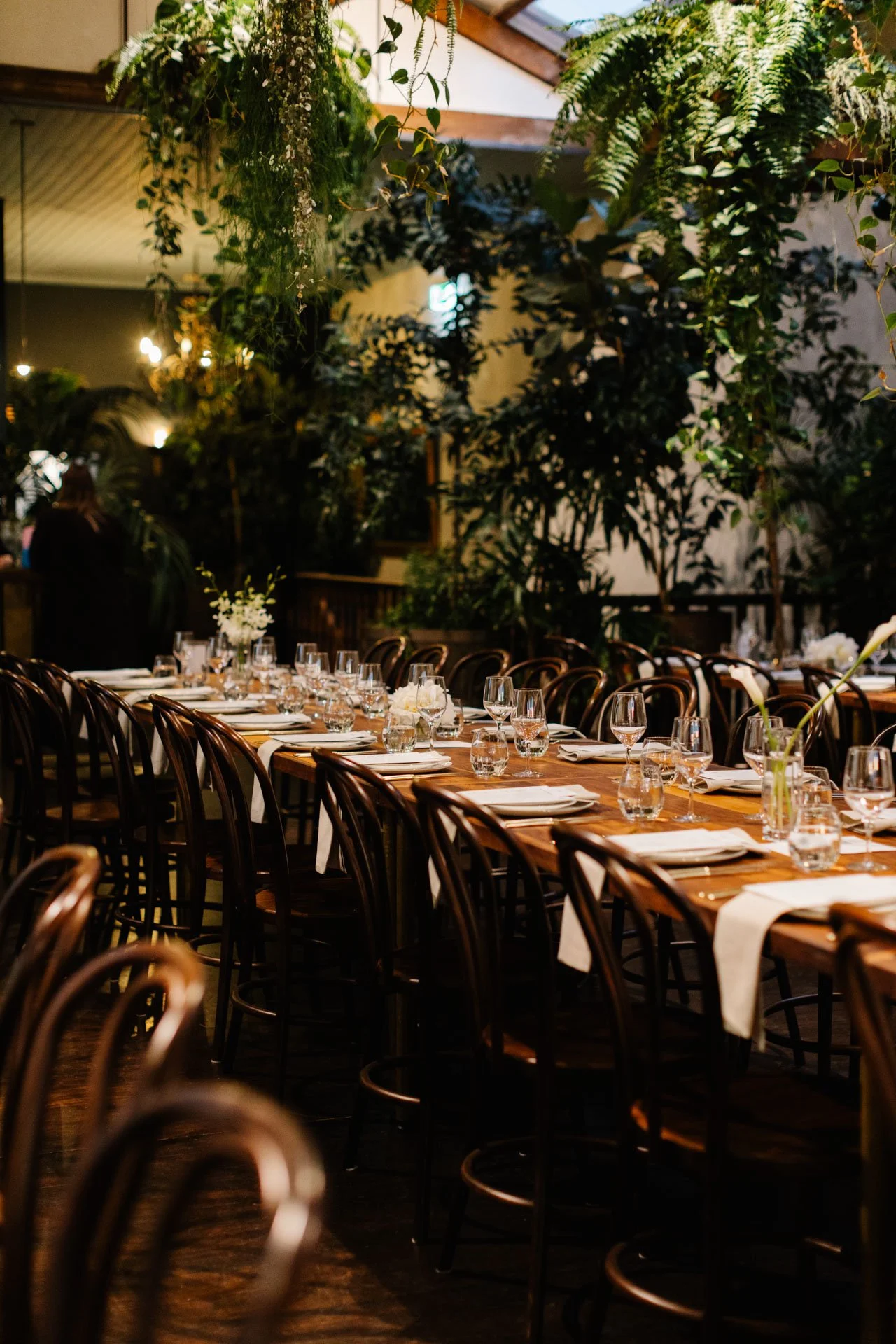 A long dining table set with white napkins, water glasses, wine glasses, and floral centerpieces in a dimly lit, elegant restaurant with hanging greenery and wood accents.