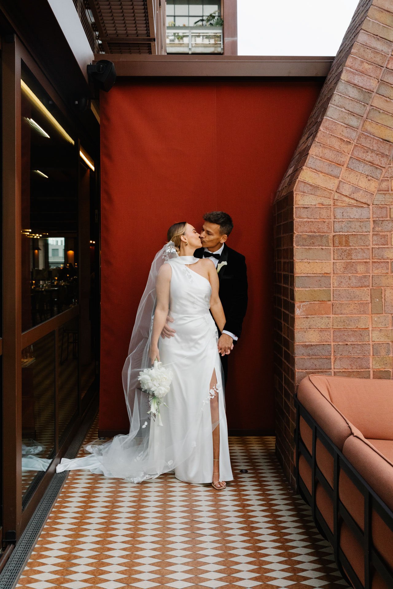 A bride and groom share a kiss indoors, standing close against a red wall. The bride wears a white gown with a veil, holding a white bouquet, and the groom wears a black tuxedo. There are brick walls and patterned floor tiles in the background.