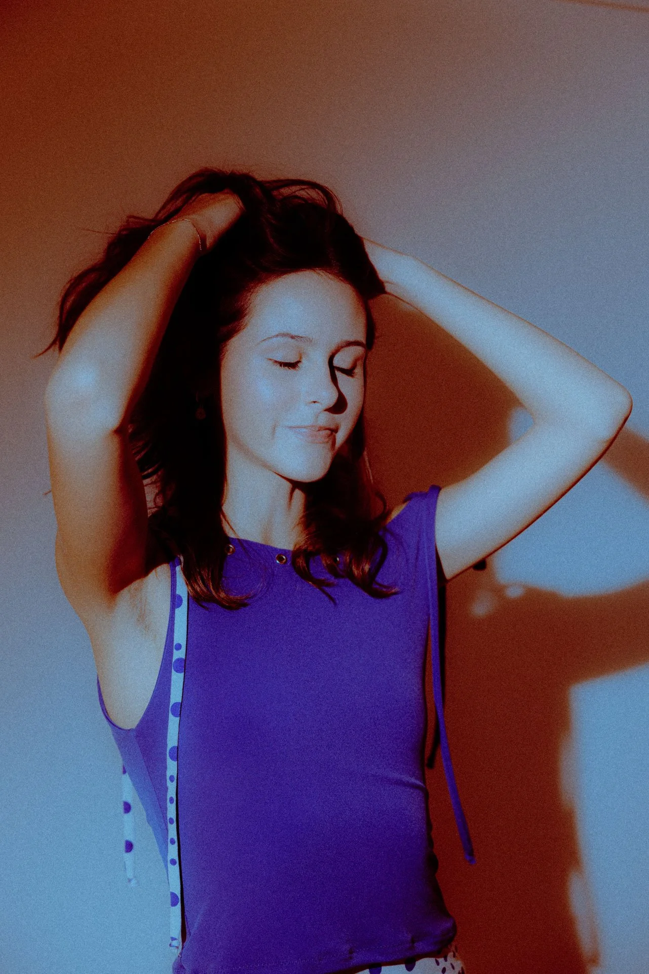 A woman with brown hair and closed eyes, wearing a purple dress with polka dot details, sitting against a wall with colorful lighting, smiling softly.