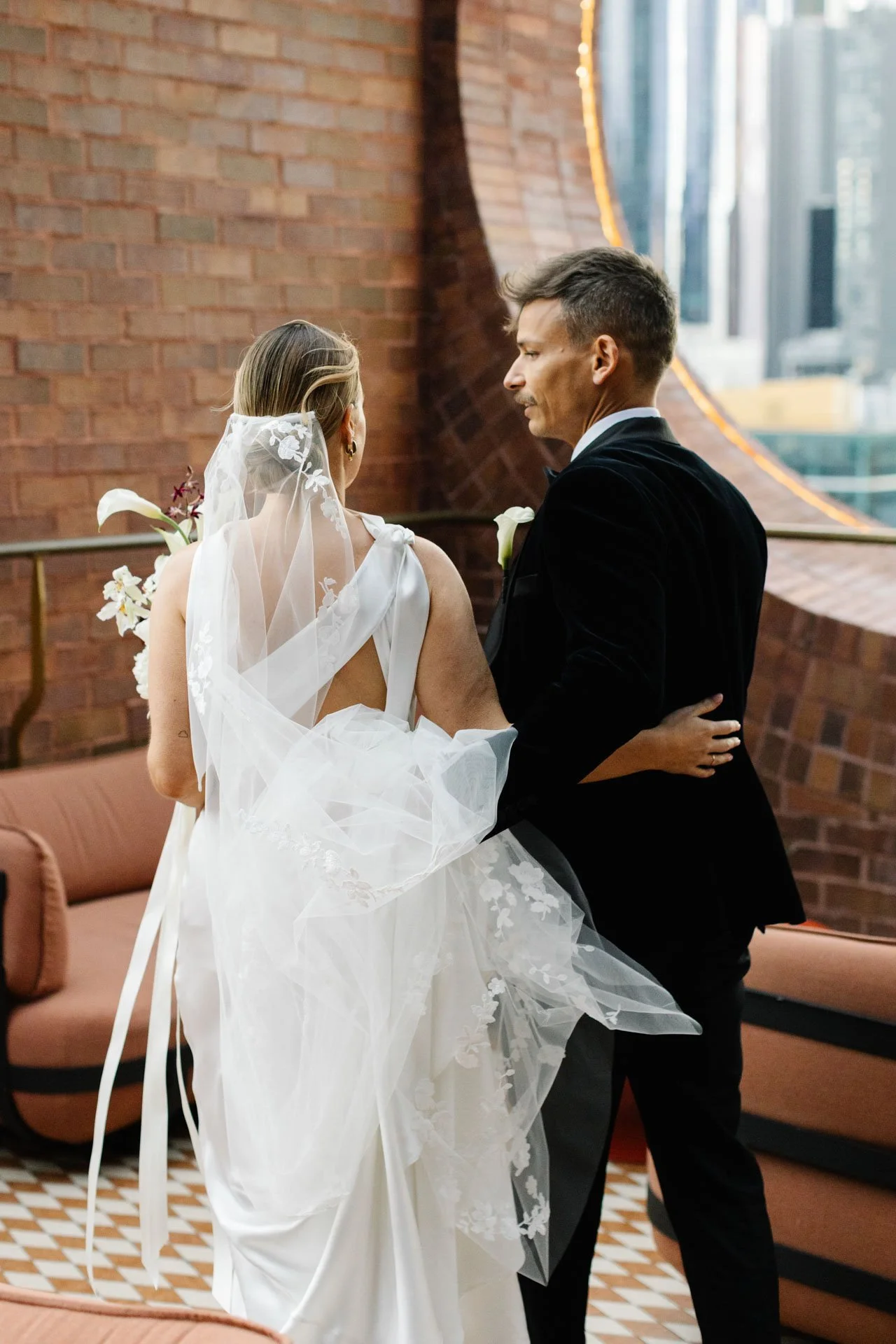 A bride and groom dressed in wedding attire sharing a moment indoors, with a brick wall and window in the background.