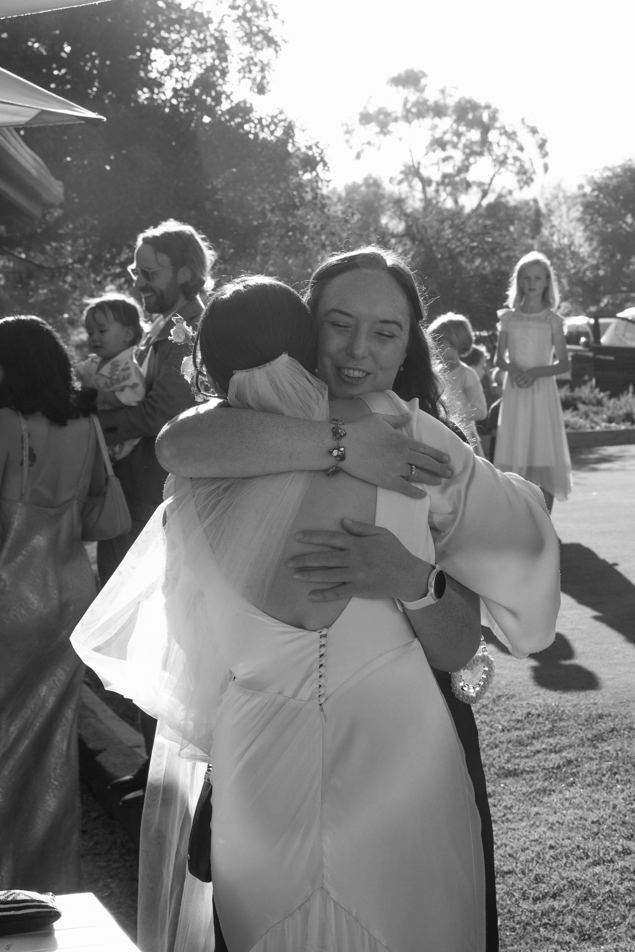 Two women, one in a wedding dress and another in a veil, hugging at an outdoor wedding reception, with guests and trees in the background.