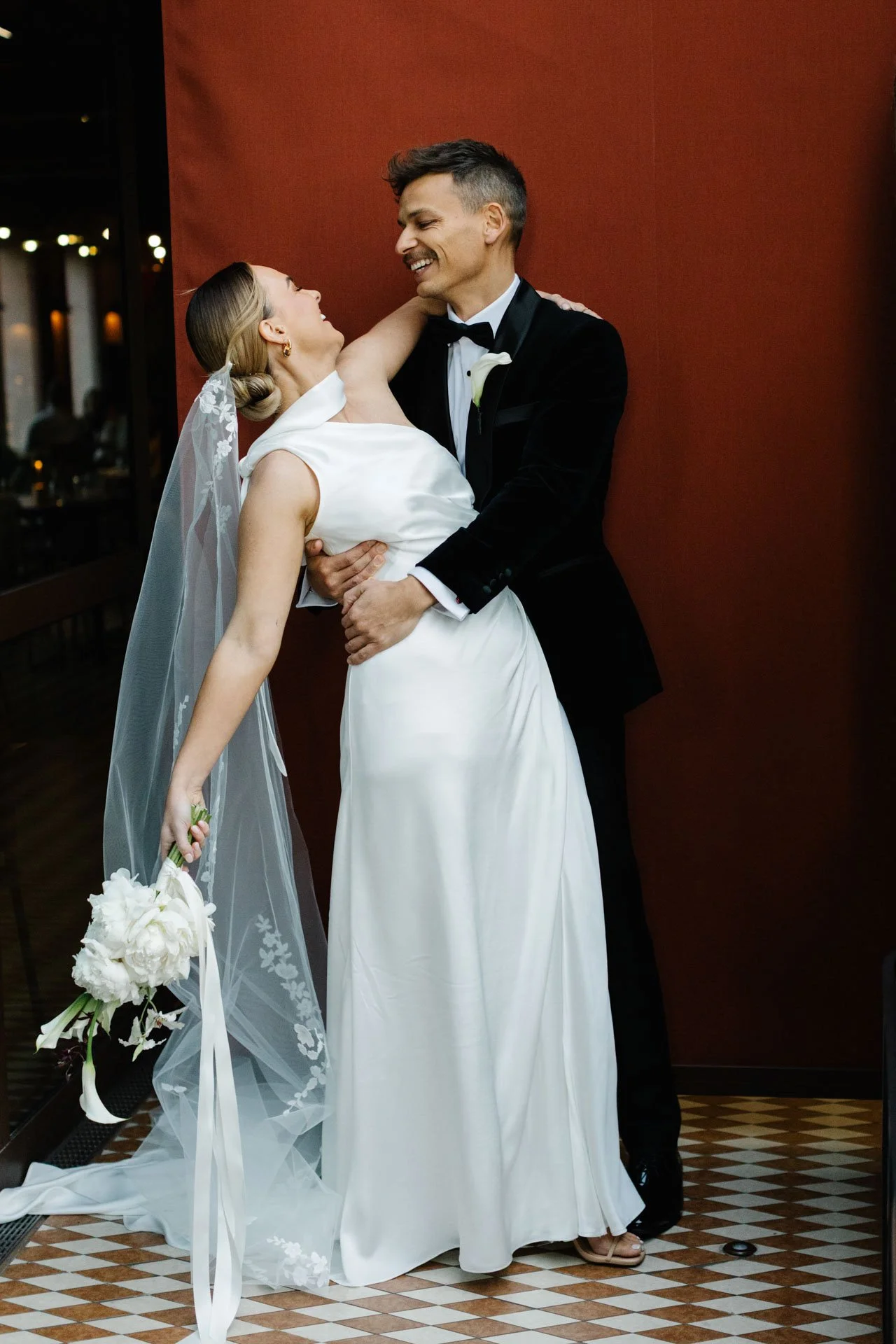A bride and groom dancing at their wedding reception, smiling at each other. The bride is wearing a wedding dress with a veil, holding a bouquet of white flowers. The groom is dressed in a black tuxedo with a bow tie, standing in front of a dark red 