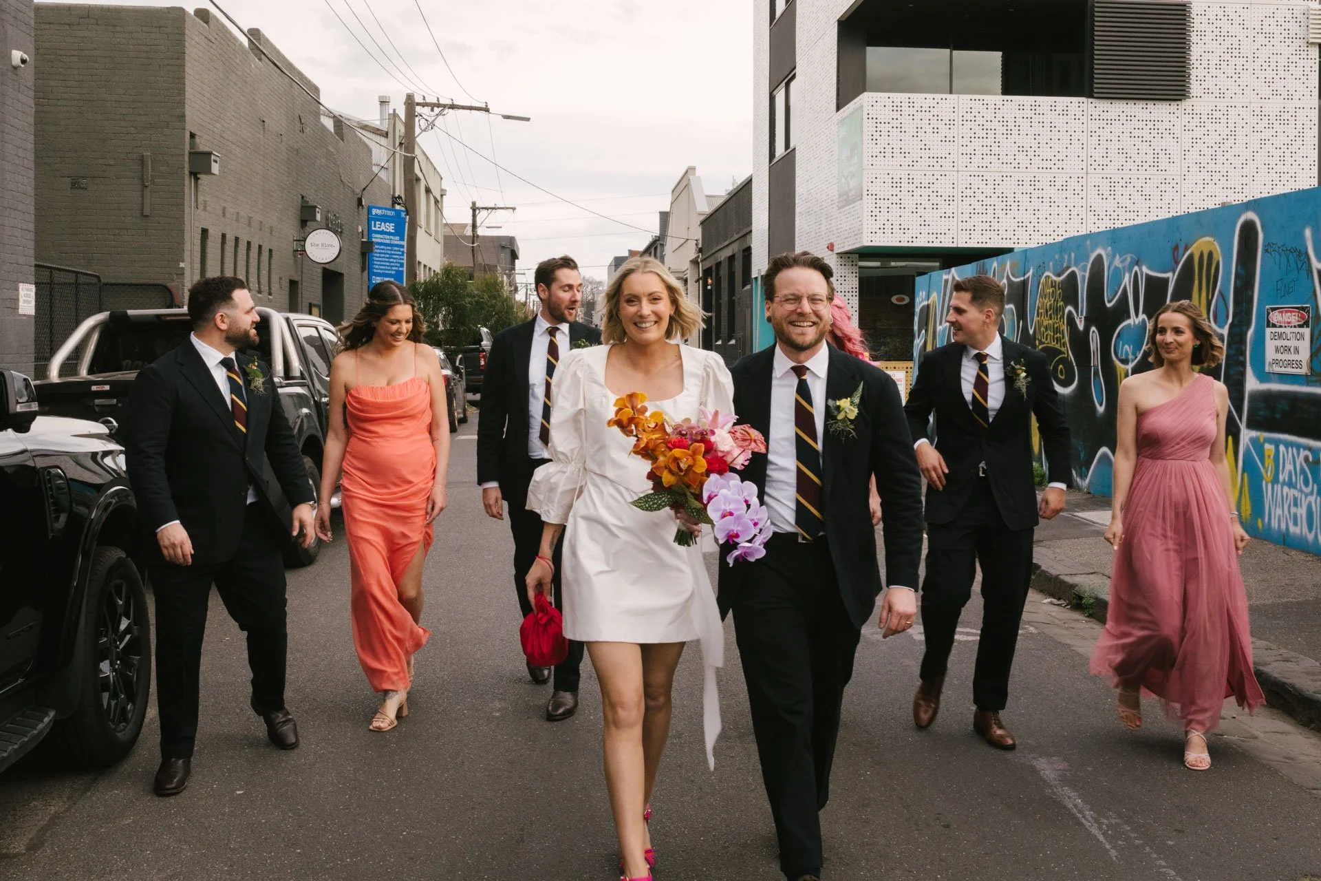 A group of people dressed in formal attire walking down a city street, with some wearing flower bouquets, suggesting a wedding celebration.