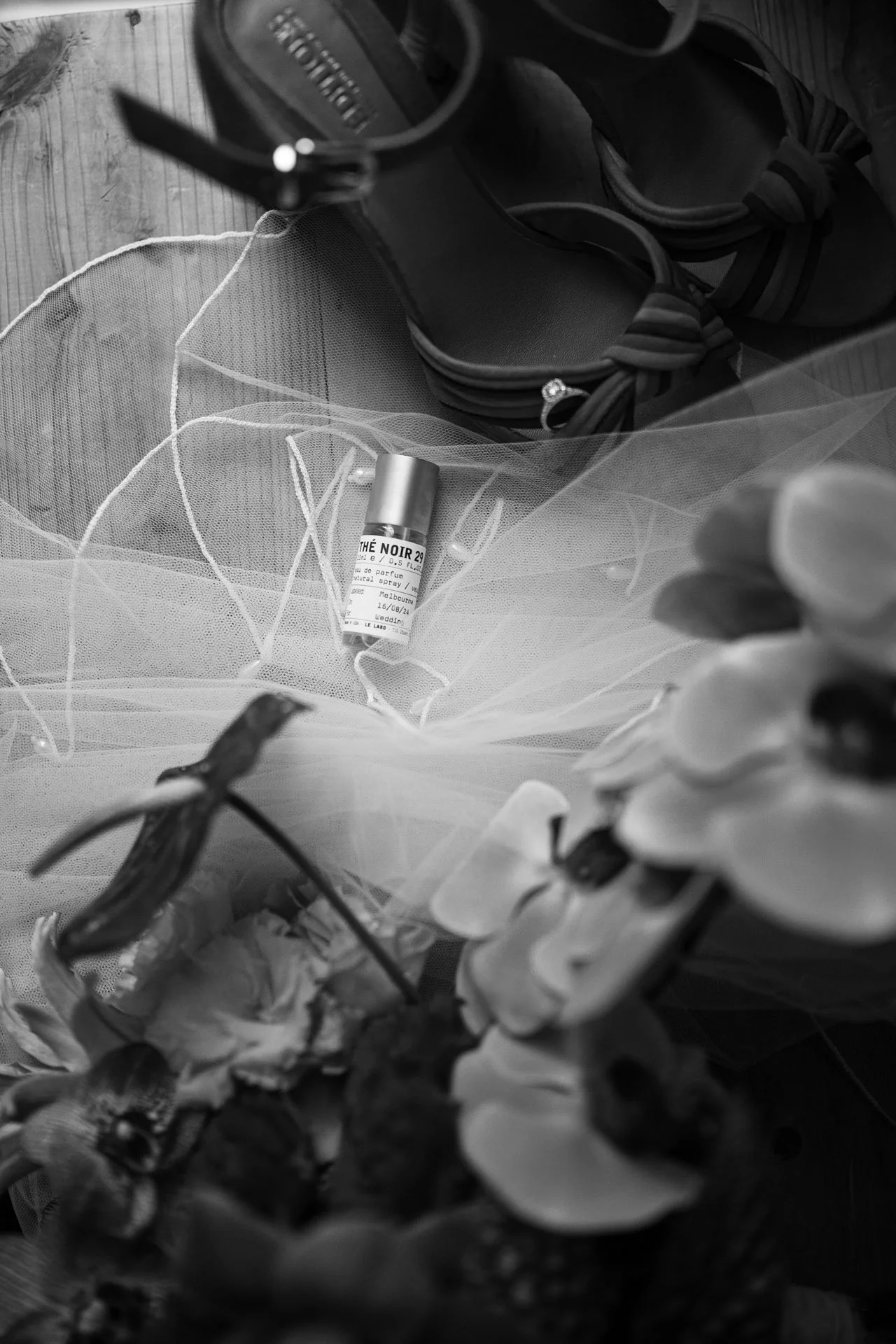 Black and white photograph of a pair of shoes, a small perfume bottle, and some fabric or netting on a wooden surface.