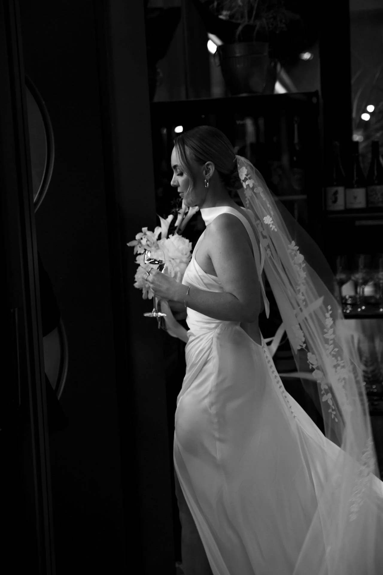 A black and white photograph of a bride in a wedding dress holding a glass of wine, standing in front of a wine fridge, with a bouquet of flowers in her other hand.