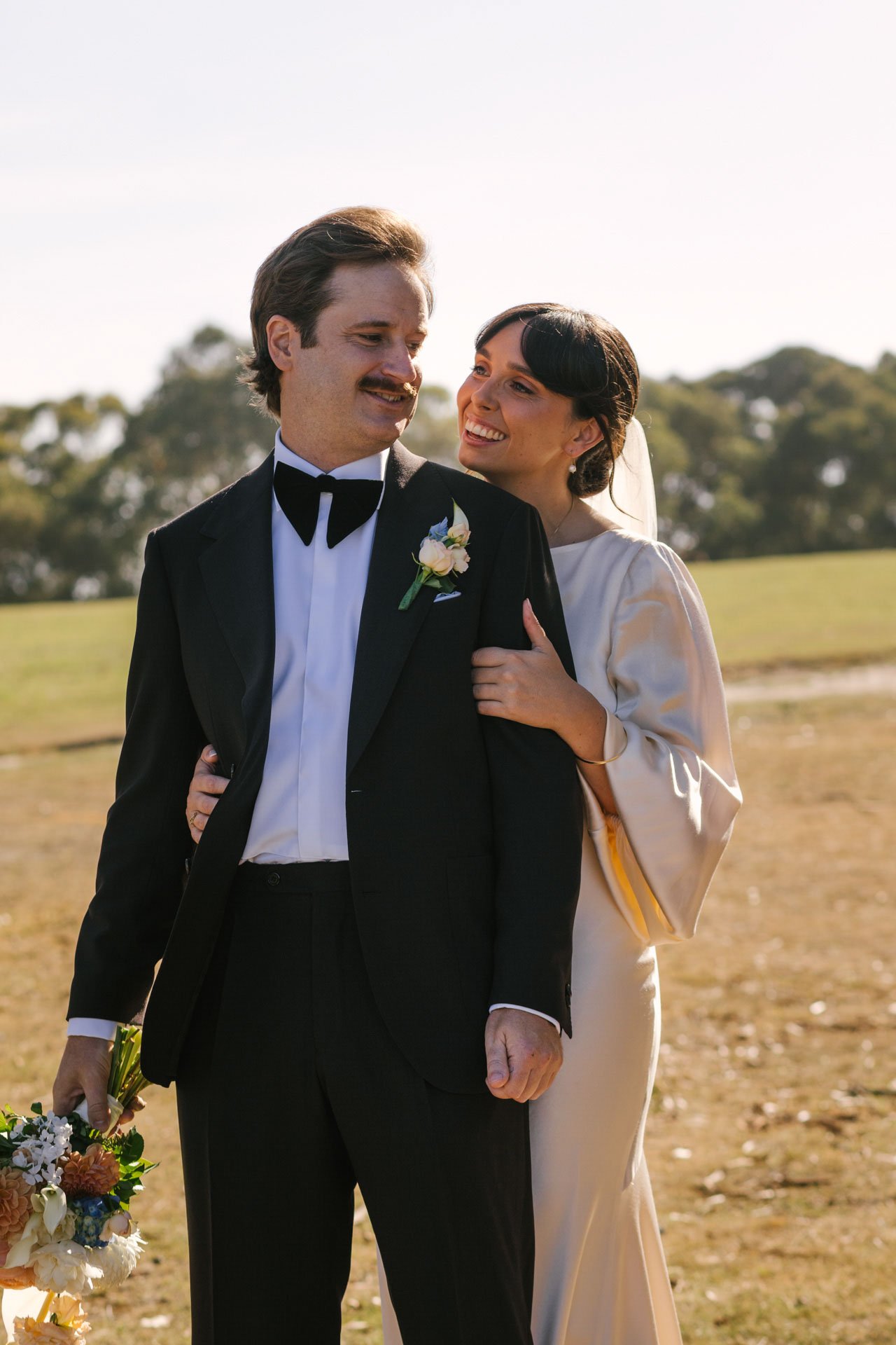 A man in a tuxedo with a boutonniere and a woman in a cream dress smiling in an outdoor setting with trees in the background.