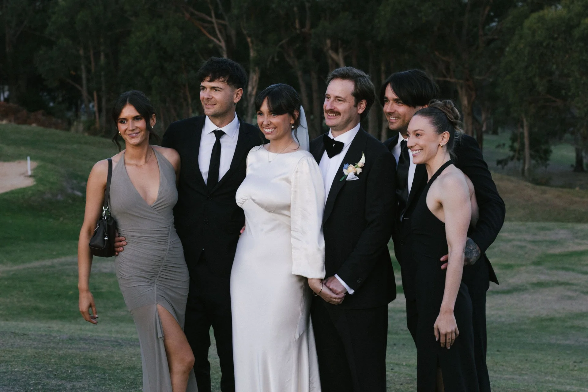 Group of six well-dressed people, including a bride and groom, standing outdoors on a green lawn with trees in the background, smiling for a photo.