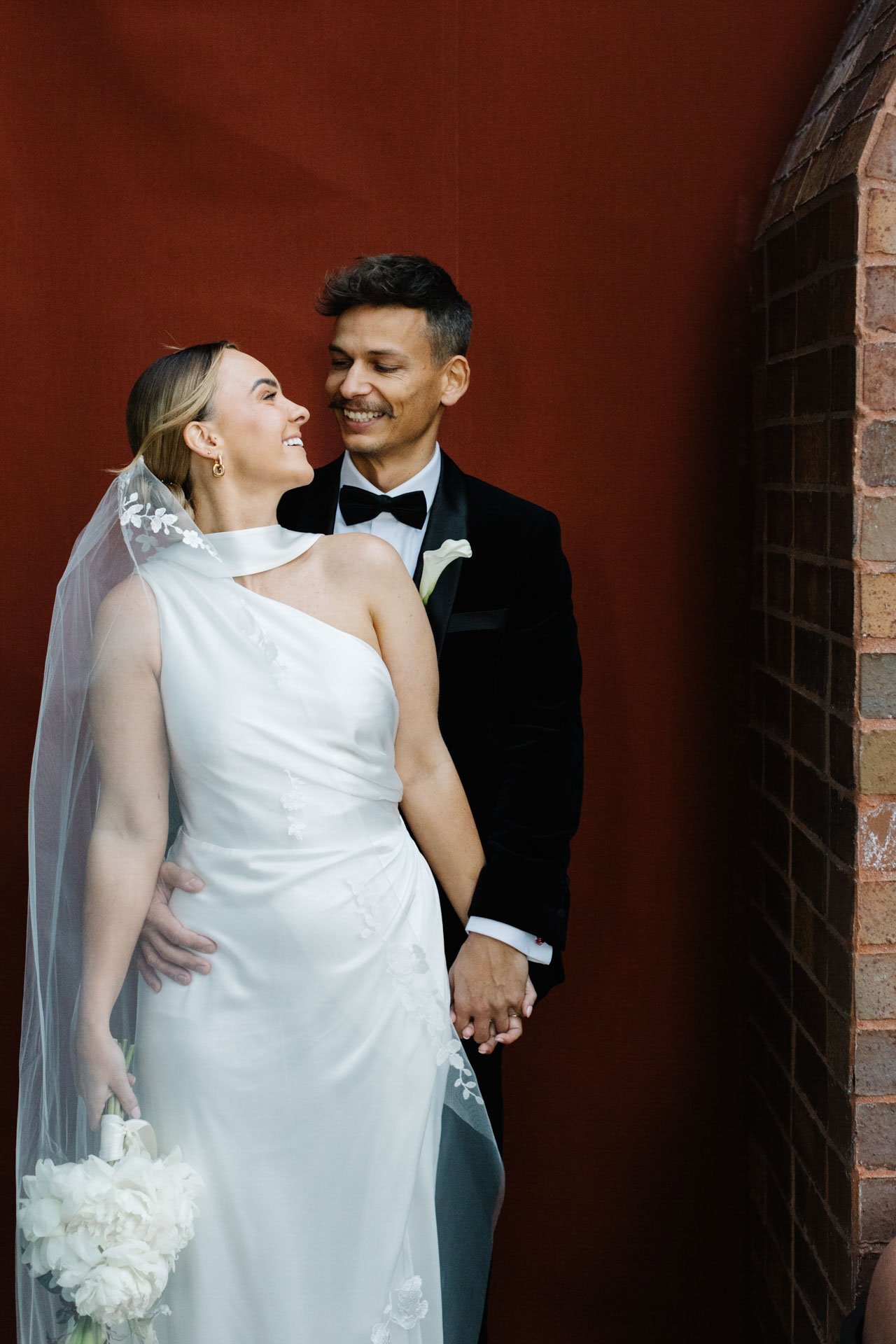 A bride and groom on their wedding day, smiling and holding hands against a red brick wall.