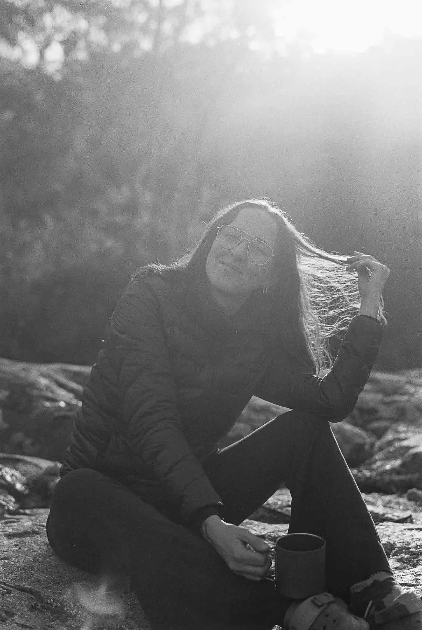 A person with long hair, wearing glasses, sitting on the ground outdoors, holding a mug, and adjusting their hair, with sunlight in the background.