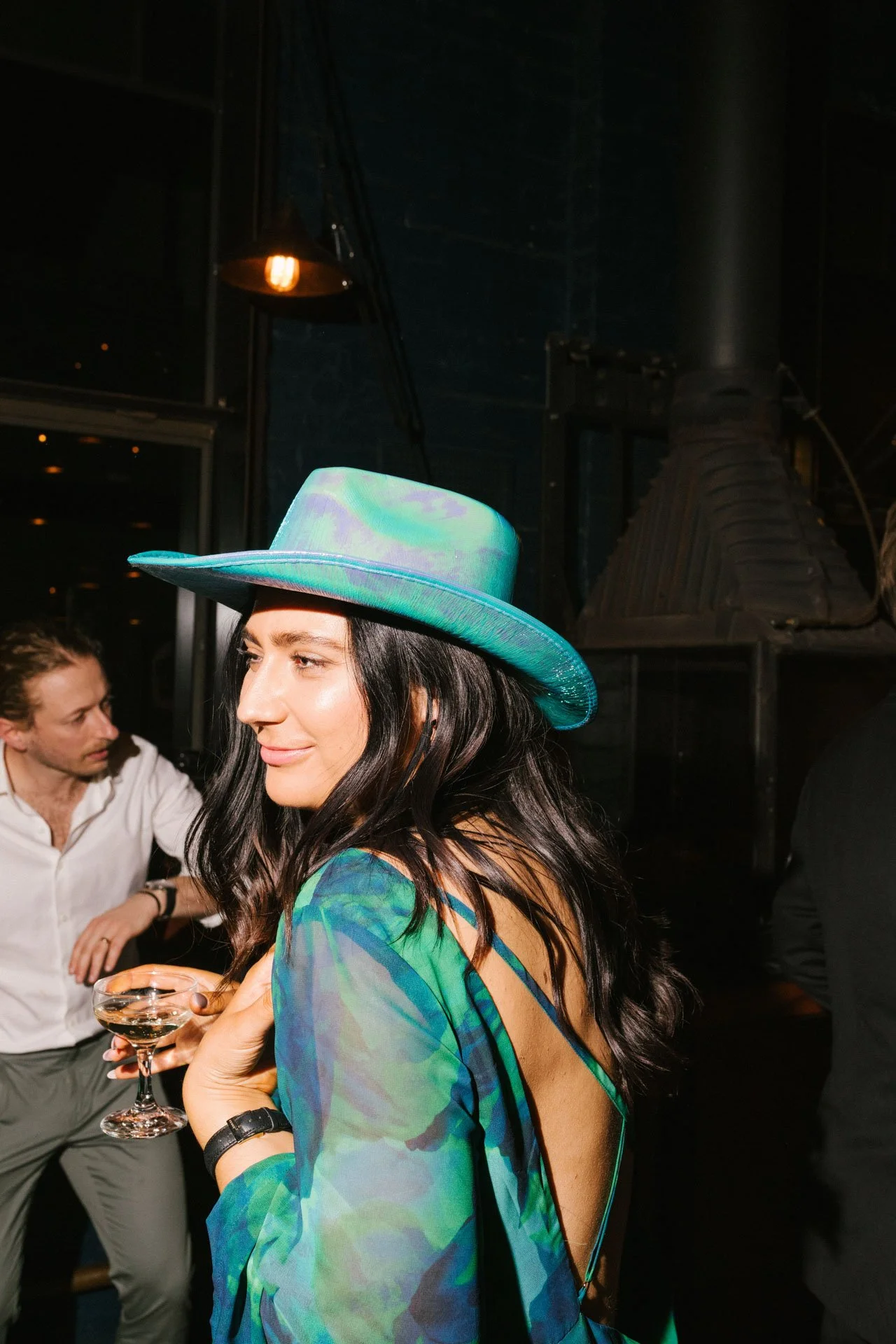 A woman with long dark hair wearing a colorful dress and a teal hat, holding a cocktail glass, at a social gathering indoors with dim lighting and other people in the background.