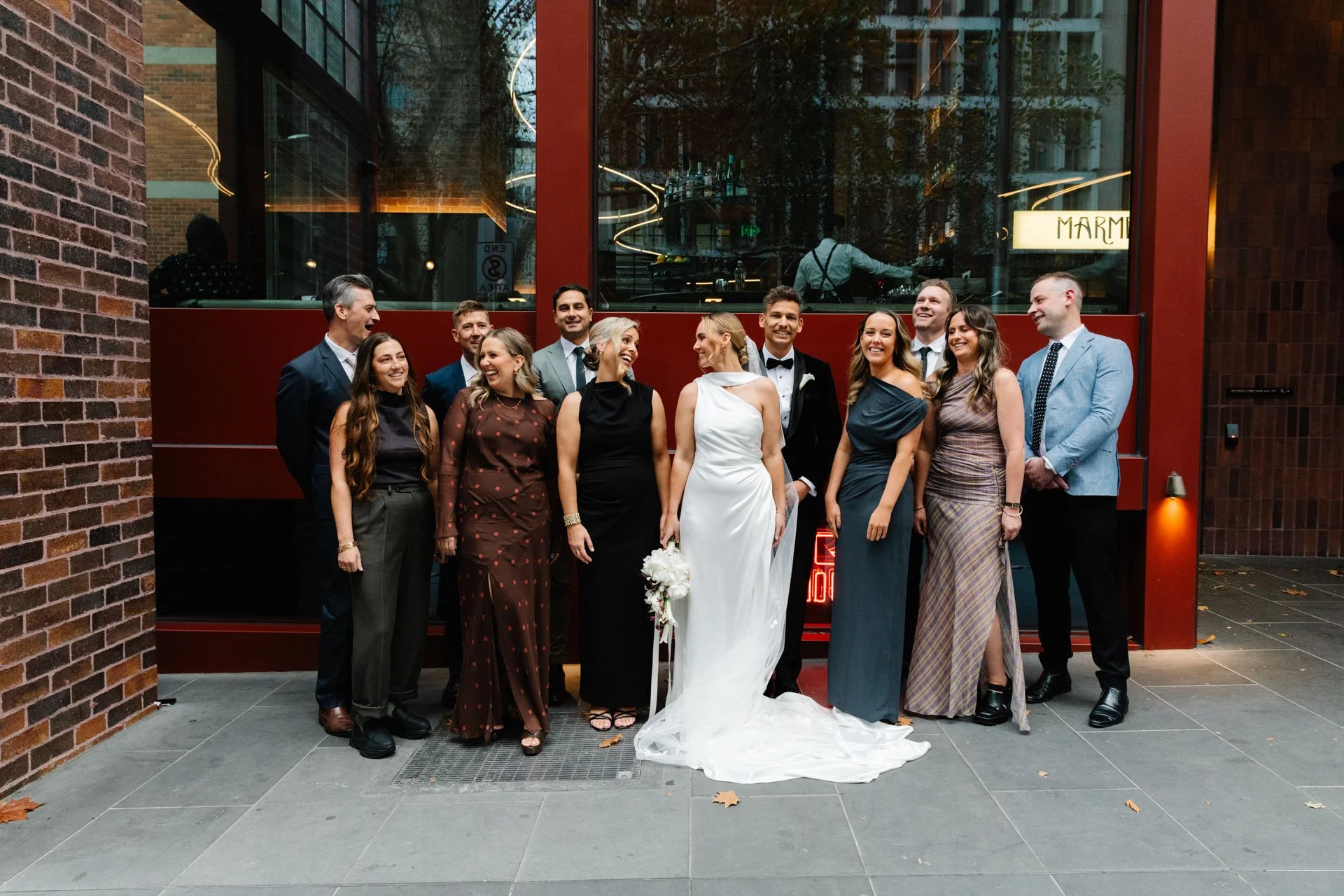 A group of people, including a bride in a white wedding dress holding a bouquet, and a groom in a tuxedo, stand together outside a building with large windows and red accents. They are smiling and posing for a photo.