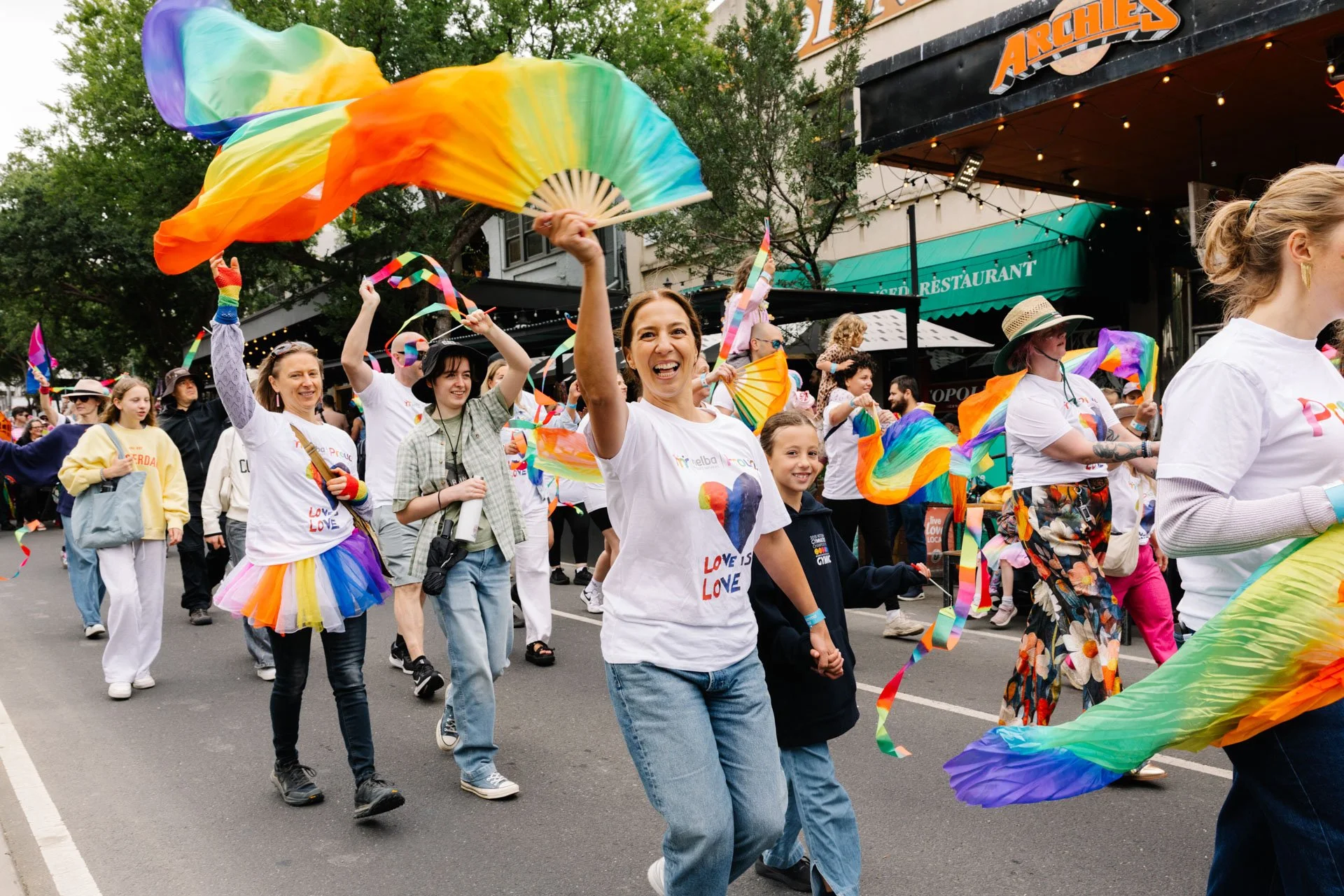 People participating in a pride parade, walking down the street, waving rainbow flags, and wearing colorful clothing. Some hold rainbow streamers and fans, celebrating diversity and love.