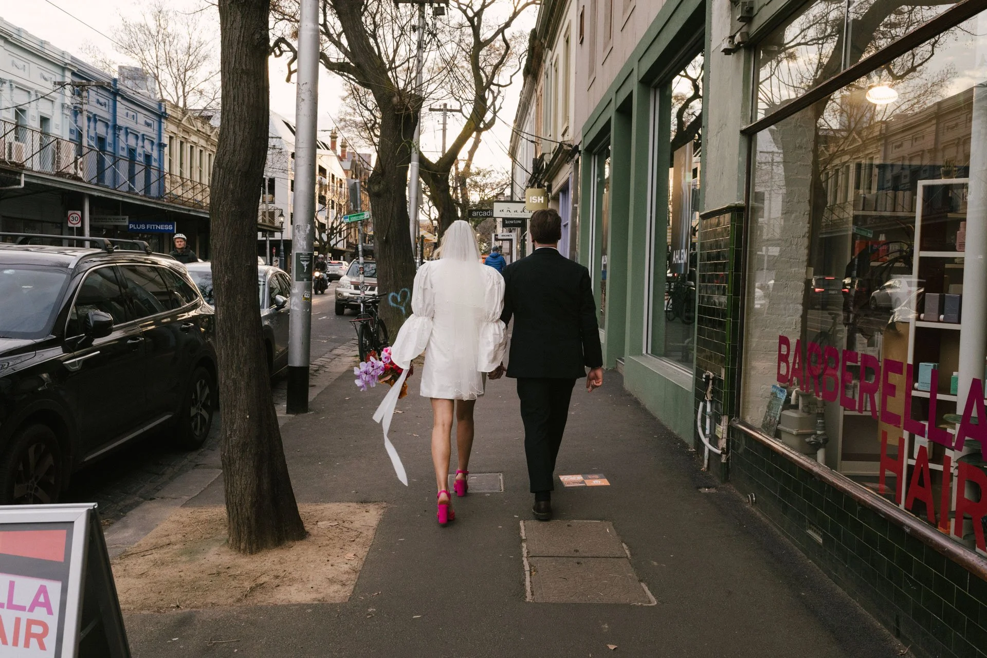 A bride in a white dress and pink heels walking hand in hand with a man in a black suit on a city sidewalk. The bride has a veil and is carrying a bouquet of pink and purple flowers. They are walking past storefronts and parked cars, with leafless tr
