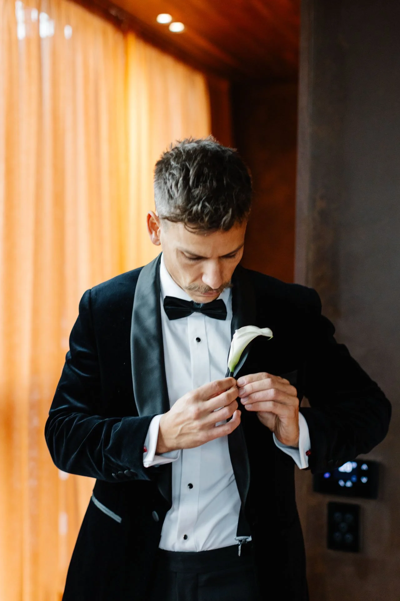 A man dressed in a black tuxedo with a white dress shirt and black bow tie is holding and preparing a white calla lily flower, looking down at it. The background has orange curtains and wooden walls.