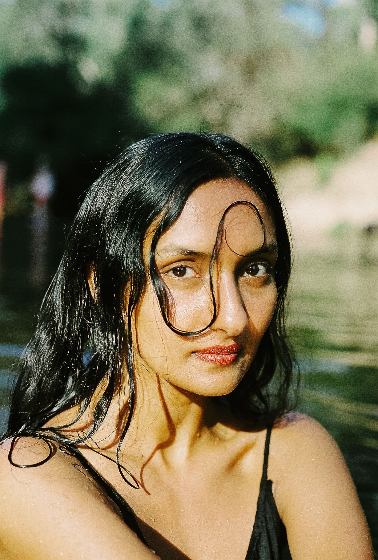 A woman with wet black hair and a curl over her face, wearing a black spaghetti strap top, standing outdoors near water with a blurred green background.