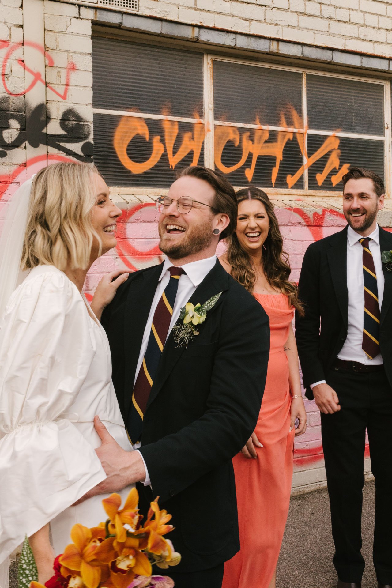 A wedding celebration with four people standing outdoors against a graffiti-covered brick wall, with the bride and groom in the foreground smiling at each other, and two guests behind them laughing.