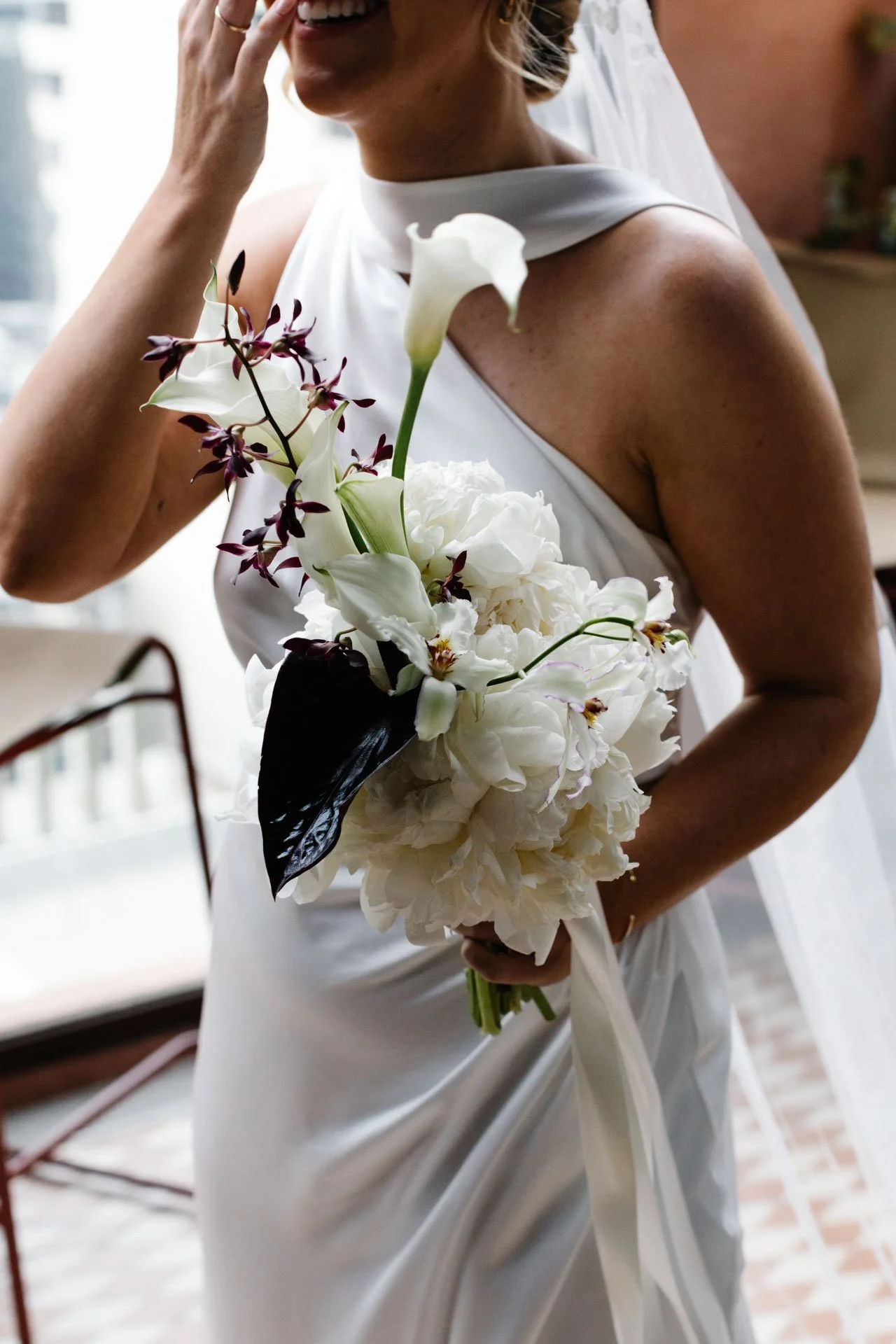 A woman in a white dress holding a bouquet of white flowers including calla lilies and peonies, smiling with her hand near her face.