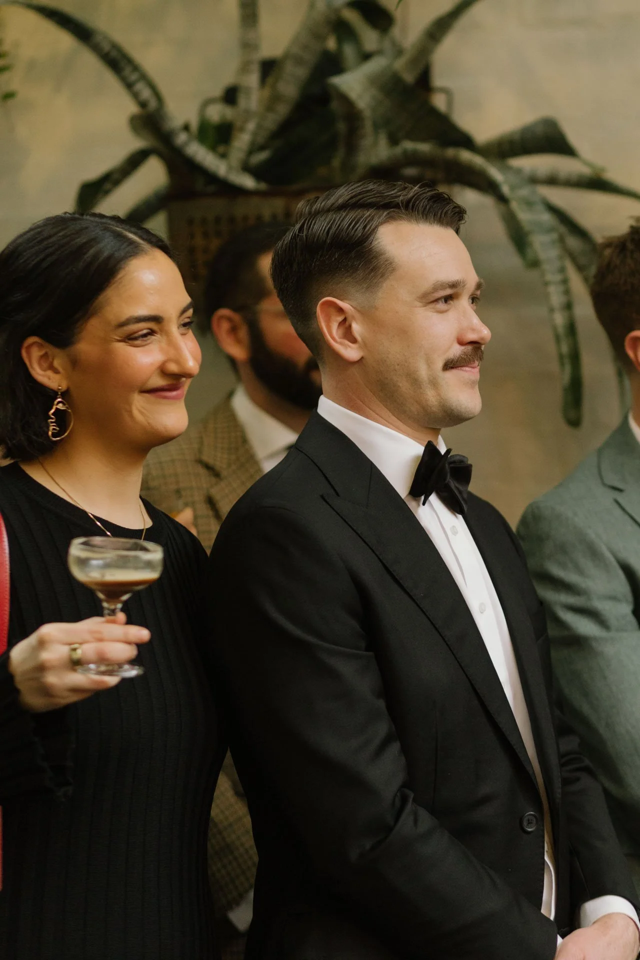 A woman in a black dress holding a cocktail glass standing next to a man in a tuxedo at a social event, with a large leafy plant in the background.
