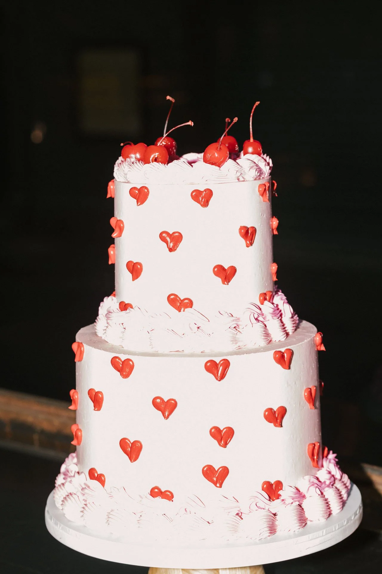 A two-tiered white wedding cake decorated with small red heart-shaped accents and topped with cherries.