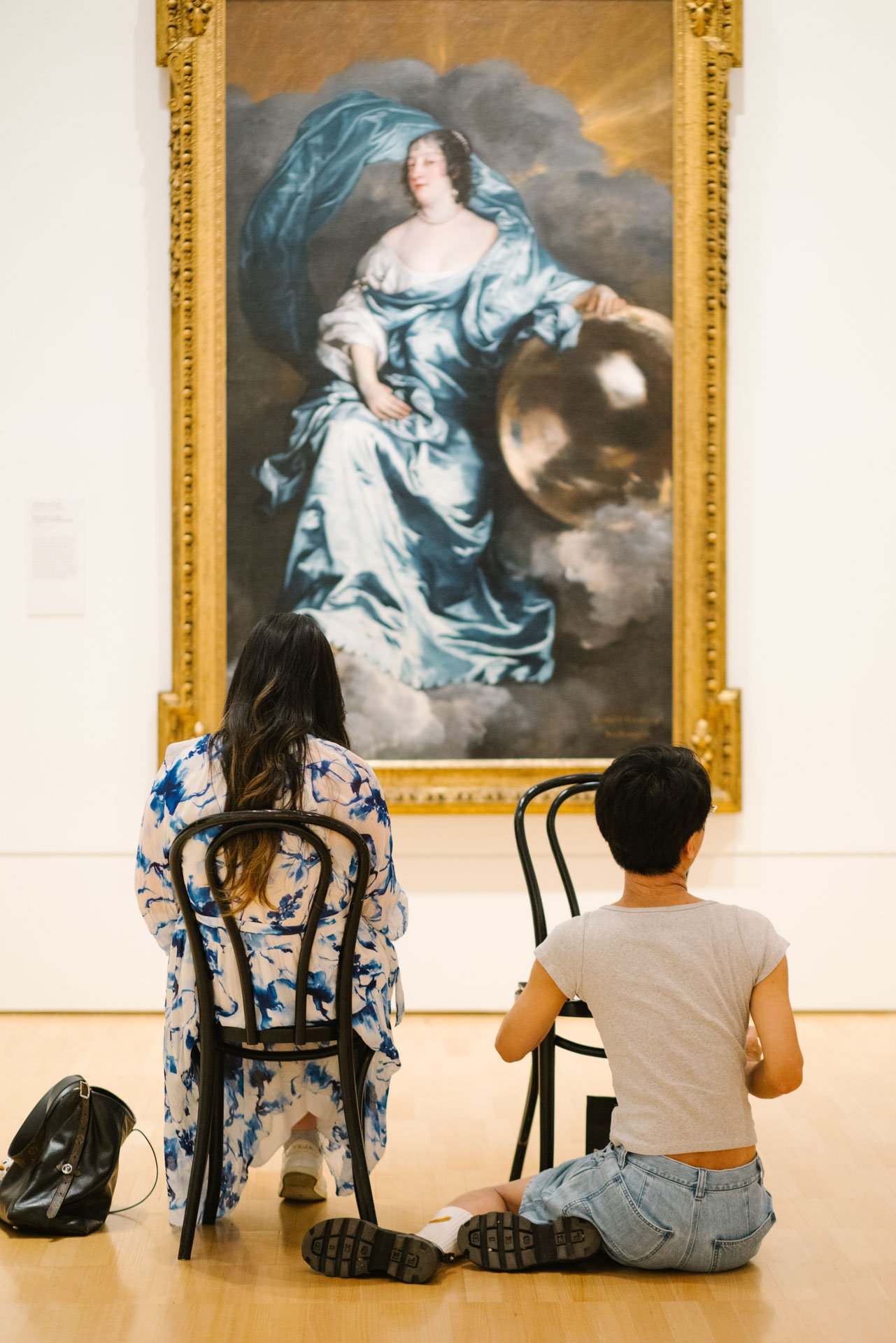 Three people in an art gallery observing a large classical painting of a woman dressed in blue, sitting with clouds in the background.