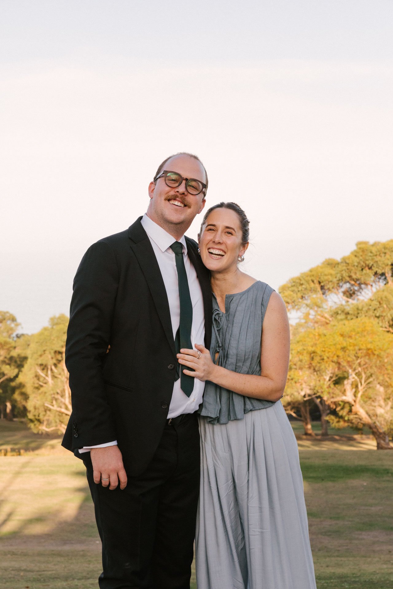 A man and woman standing together outdoors during sunset, smiling, with trees in the background.