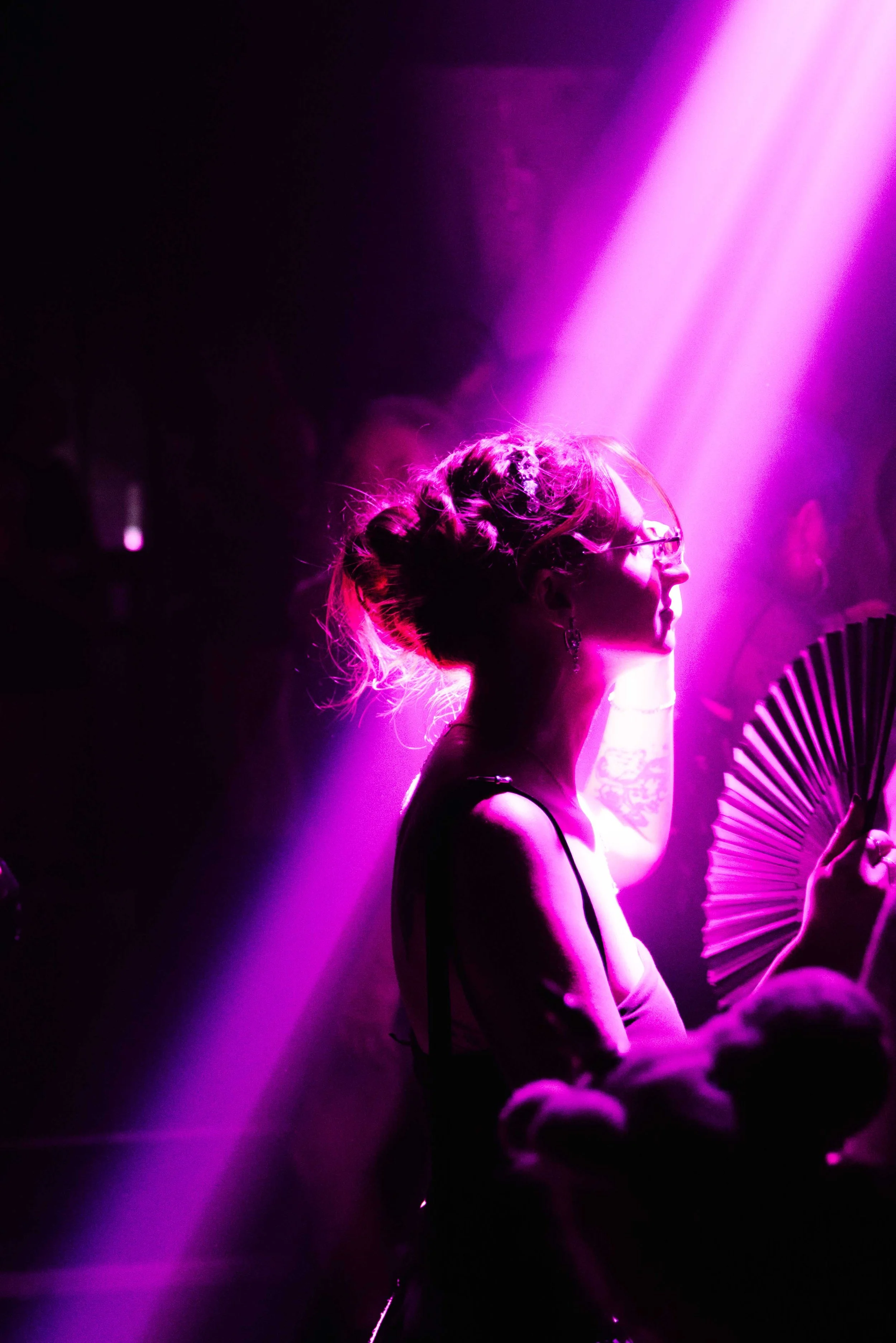 A woman with glasses and her hair in a messy bun is illuminated by pink and purple stage lighting, holding a pink paper fan, in a dark setting with other faint figures in the background.