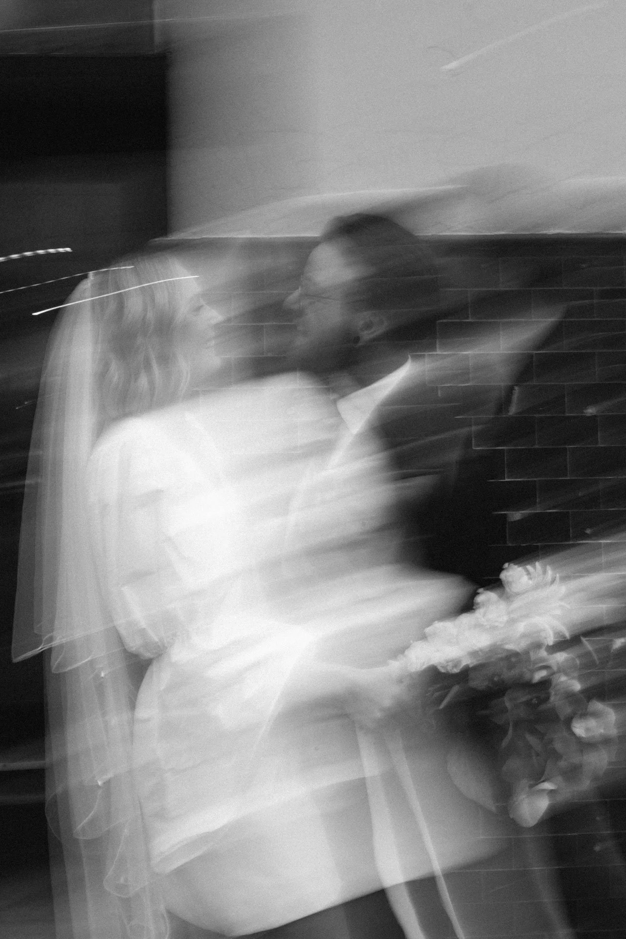 A blurred black and white photo of a person with glasses in a tuxedo, holding a bouquet of flowers, with a woman in a light-colored dress also holding flowers, against a brick wall background.